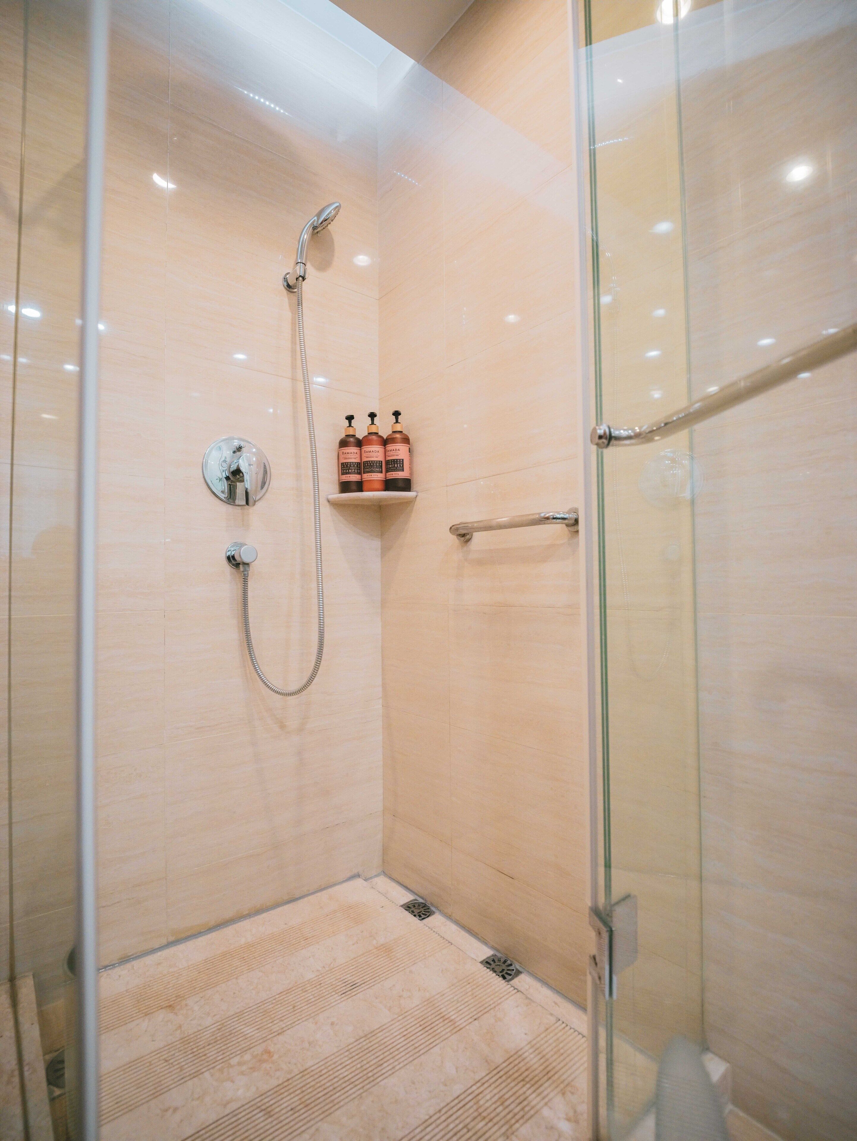 A glass-enclosed shower with a handheld showerhead and a wall-mounted faucet. There is a shelf with two bottles of shampoo and conditioner.