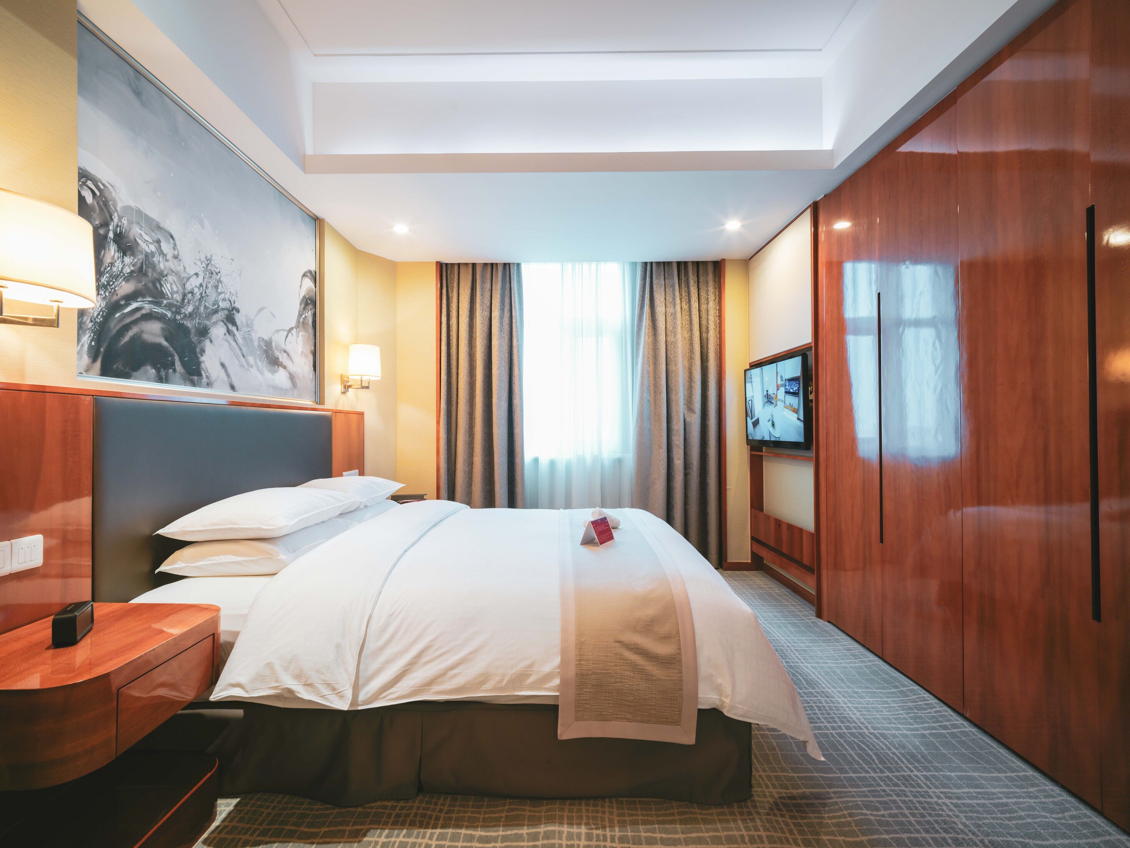 A hotel room with a bed, a desk, a TV, and a painting on the wall.