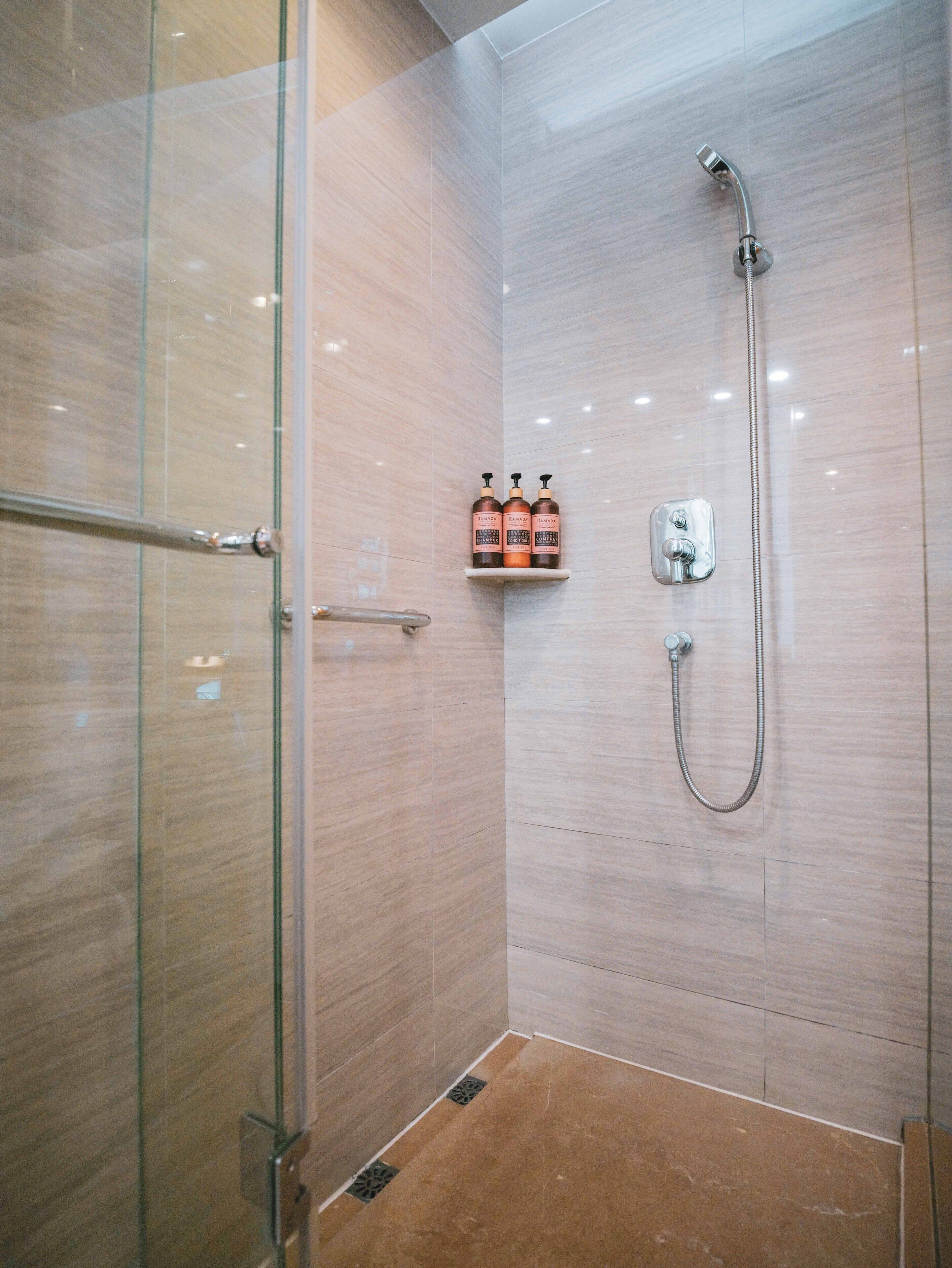A modern shower with a glass door, a handheld showerhead, and a shelf with three bottles of shampoo.