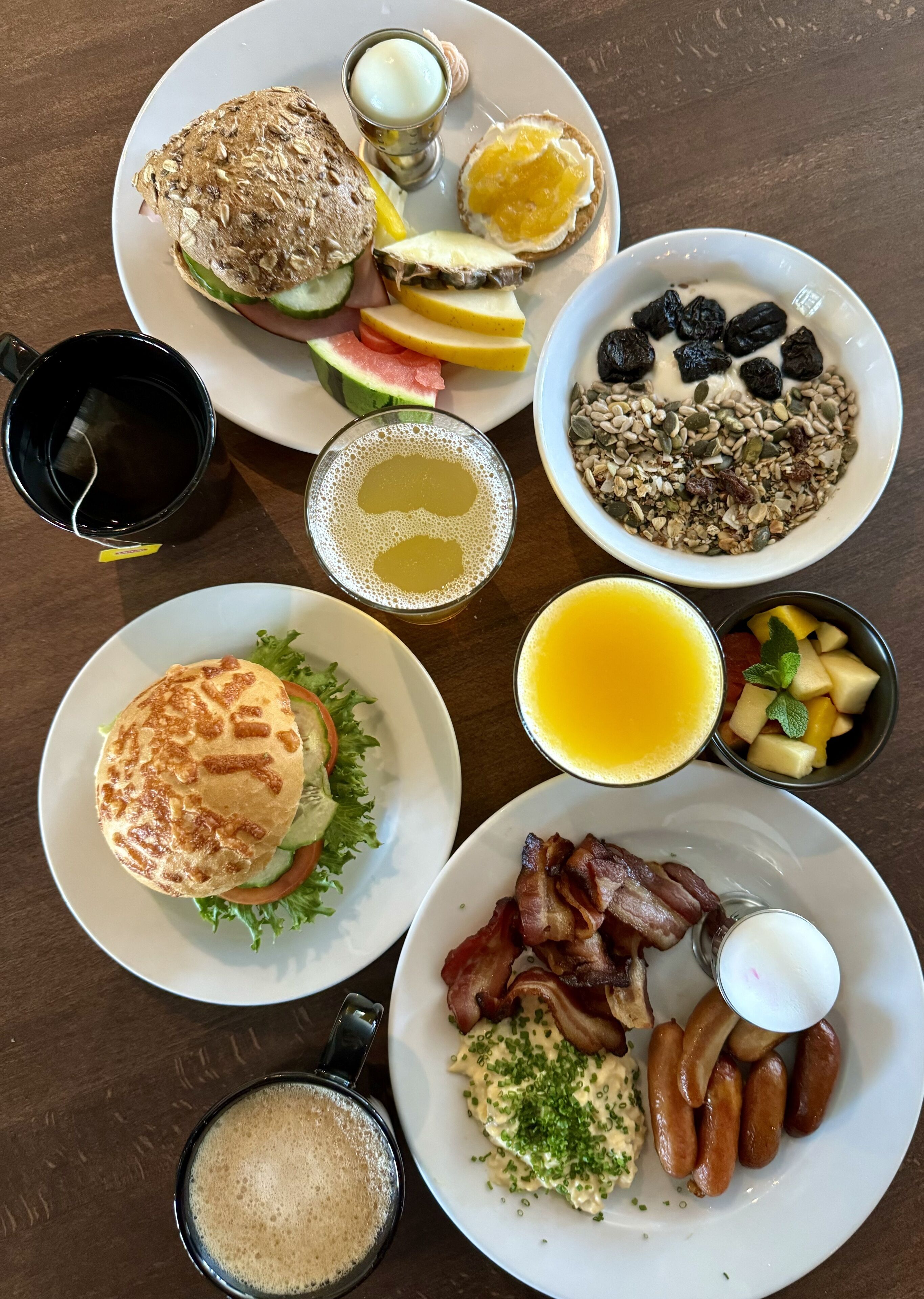 A breakfast spread with various dishes including a sandwich, fruit, eggs, bacon, sausages, and a bowl of cereal.