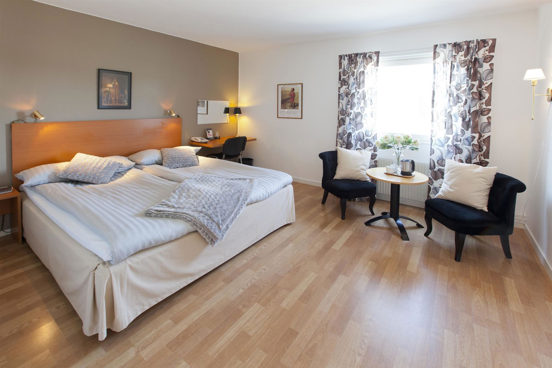 A hotel room with a large bed, two chairs, a small table, a desk with a chair, and a window with curtains.