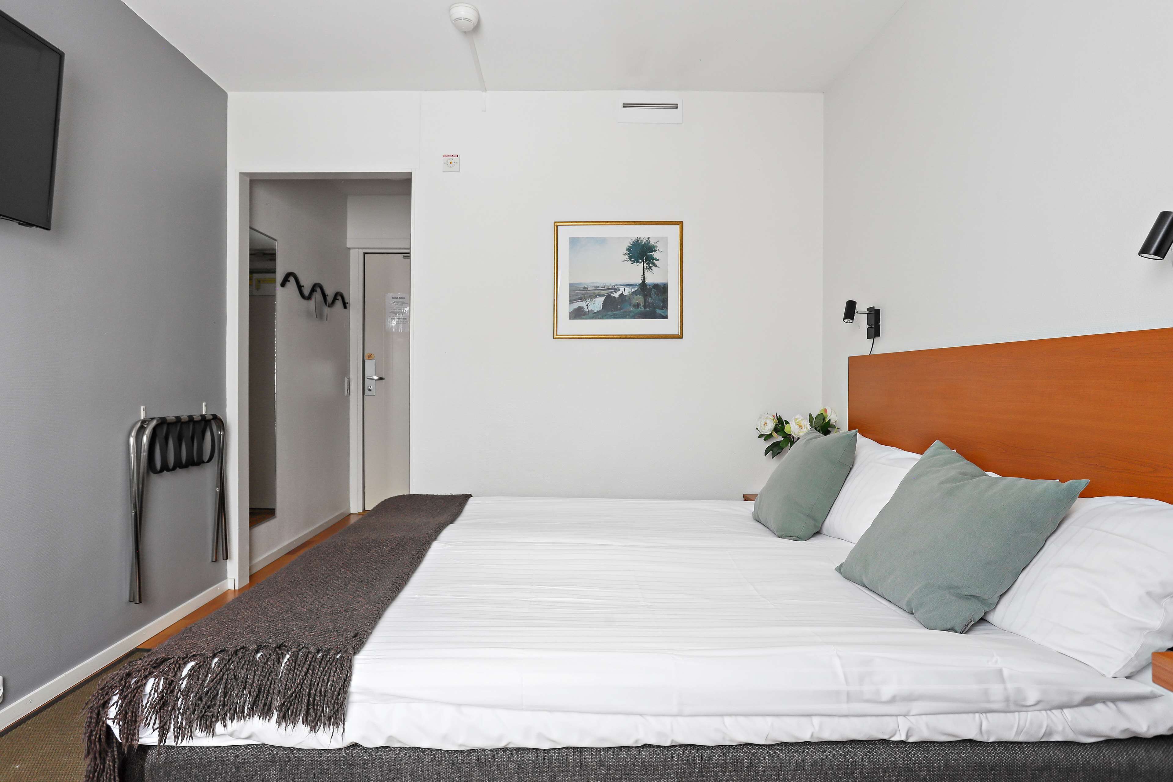 A modern hotel room with a neatly made bed, a framed picture on the wall, and a wooden headboard.