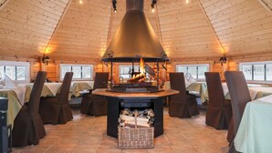 A restaurant with a central wood-burning stove, tables with brown chairs, and a high wooden ceiling.