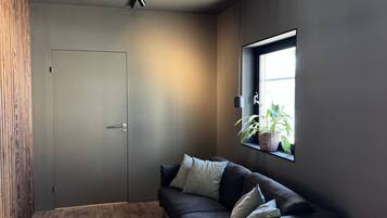 A modern room with a dark grey sofa, a small round table with glasses, and a window with plants.
