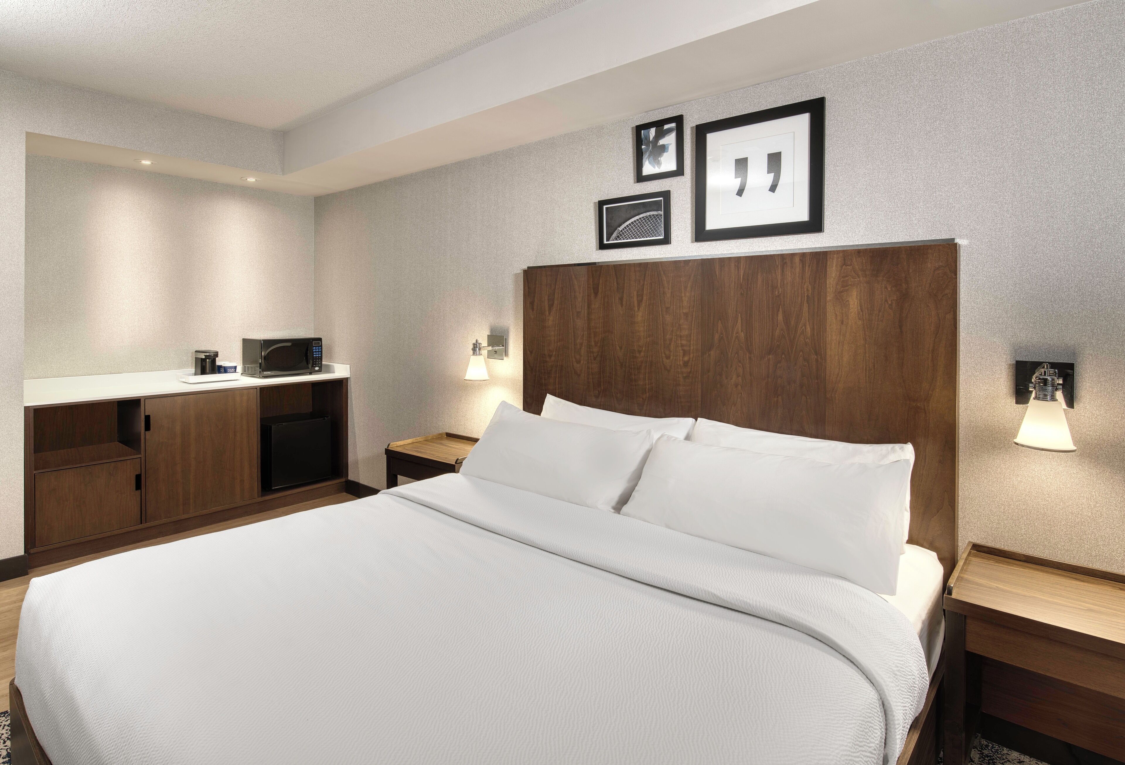 Une chambre d’hôtel avec un grand lit, une tête de lit en bois, une table de chevet avec une lampe, un petit bureau avec un micro-ondes et des tableaux encadrés accrochés au mur.