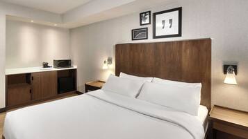 Une chambre d’hôtel avec un grand lit, une tête de lit en bois, une table de chevet avec une lampe, un petit bureau avec un micro-ondes et des tableaux encadrés accrochés au mur.