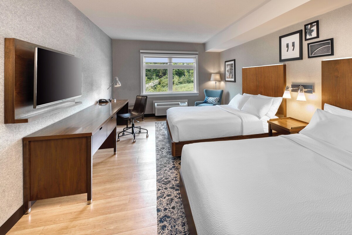 A hotel room with two beds, a desk, a TV, and a window with a view of greenery.
