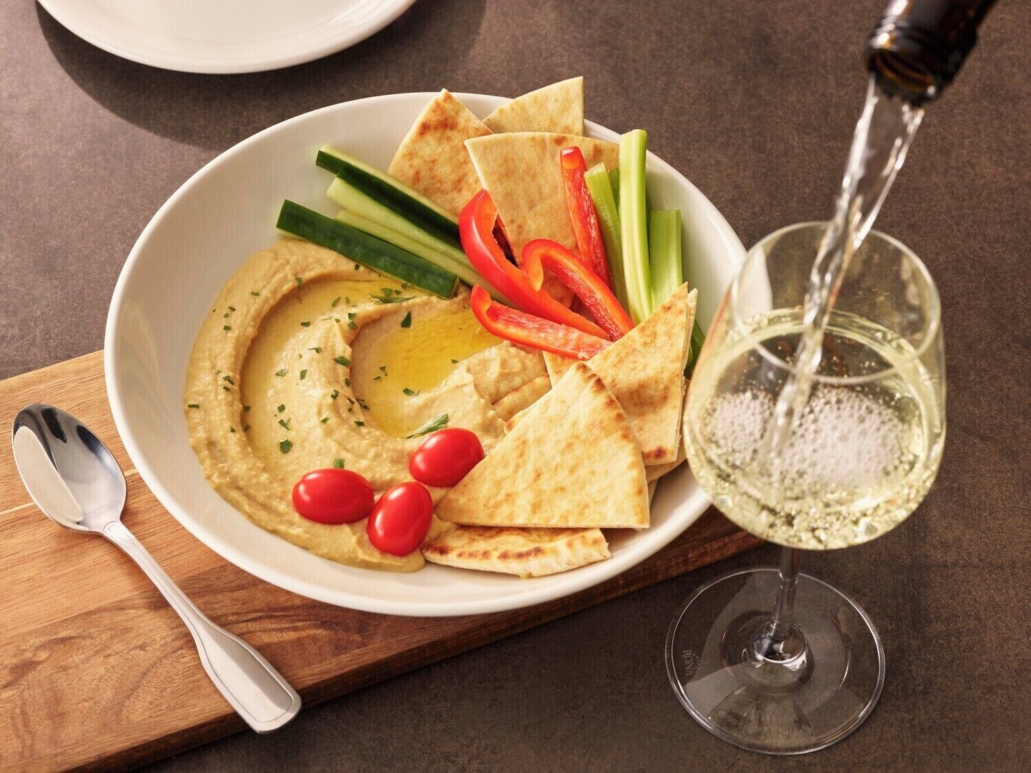 A bowl of hummus with pita chips, vegetables, and a glass of water on a wooden board.
