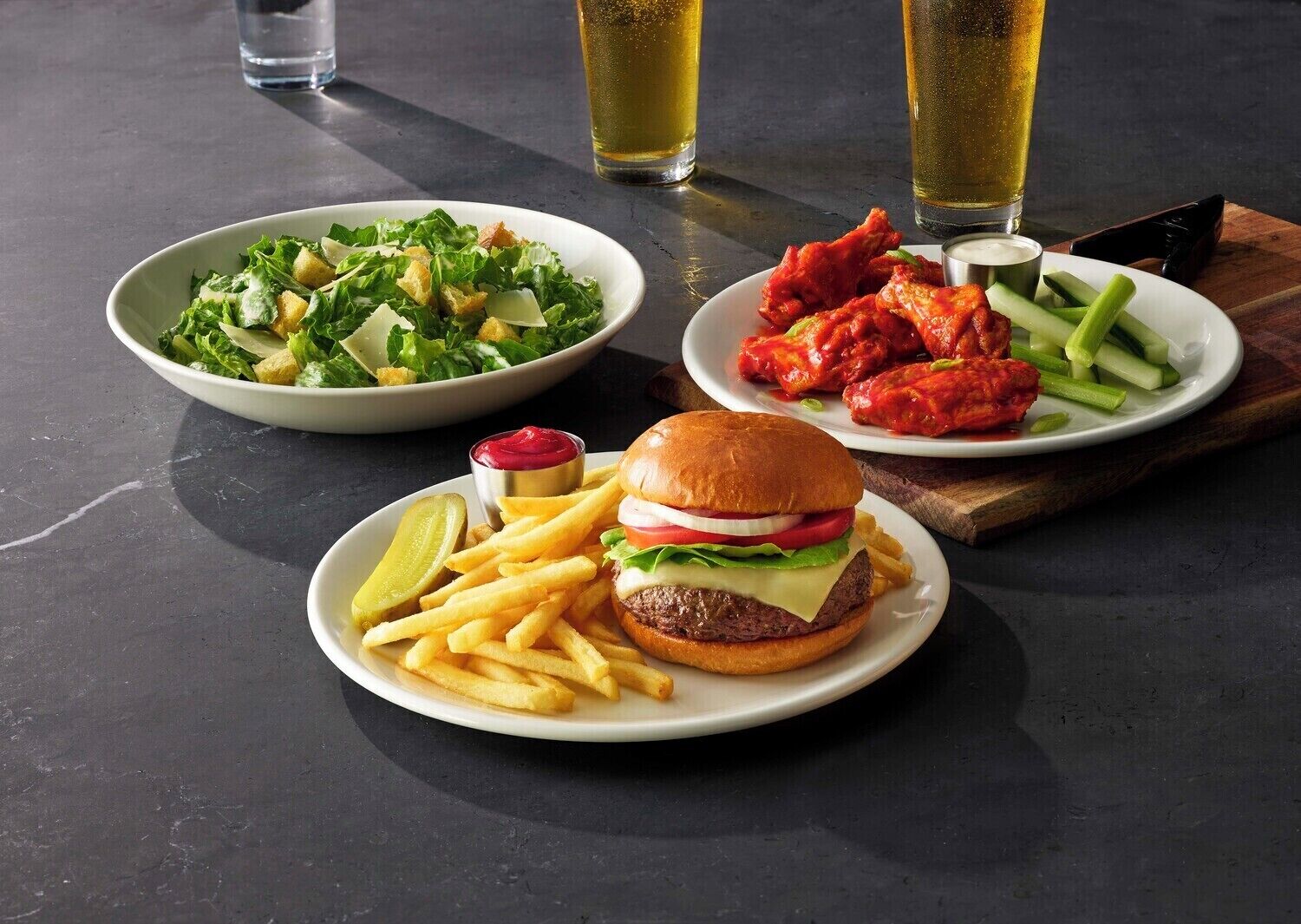 Un plato con hamburguesa, papas fritas, ensalada y alitas de pollo, con bebidas al fondo.