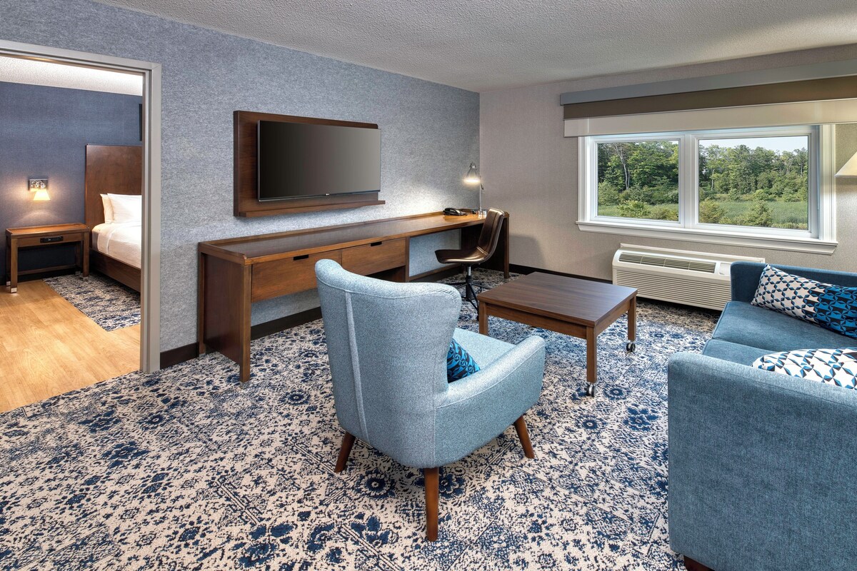 A modern hotel room with a large bed, a desk with a flat-screen TV, a comfortable seating area with two sofas, and a large window offering a view of the outdoors.