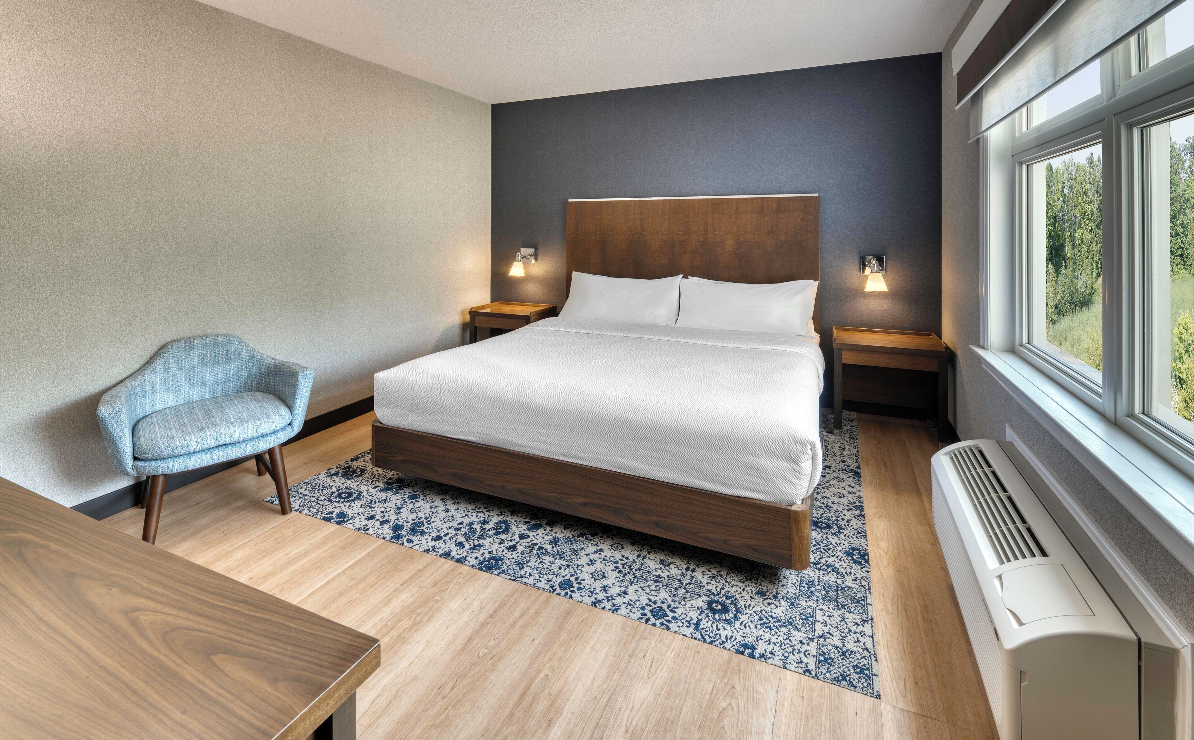 Une chambre moderne avec un grand lit, une chaise, une table de chevet et une fenêtre donnant sur un paysage verdoyant.