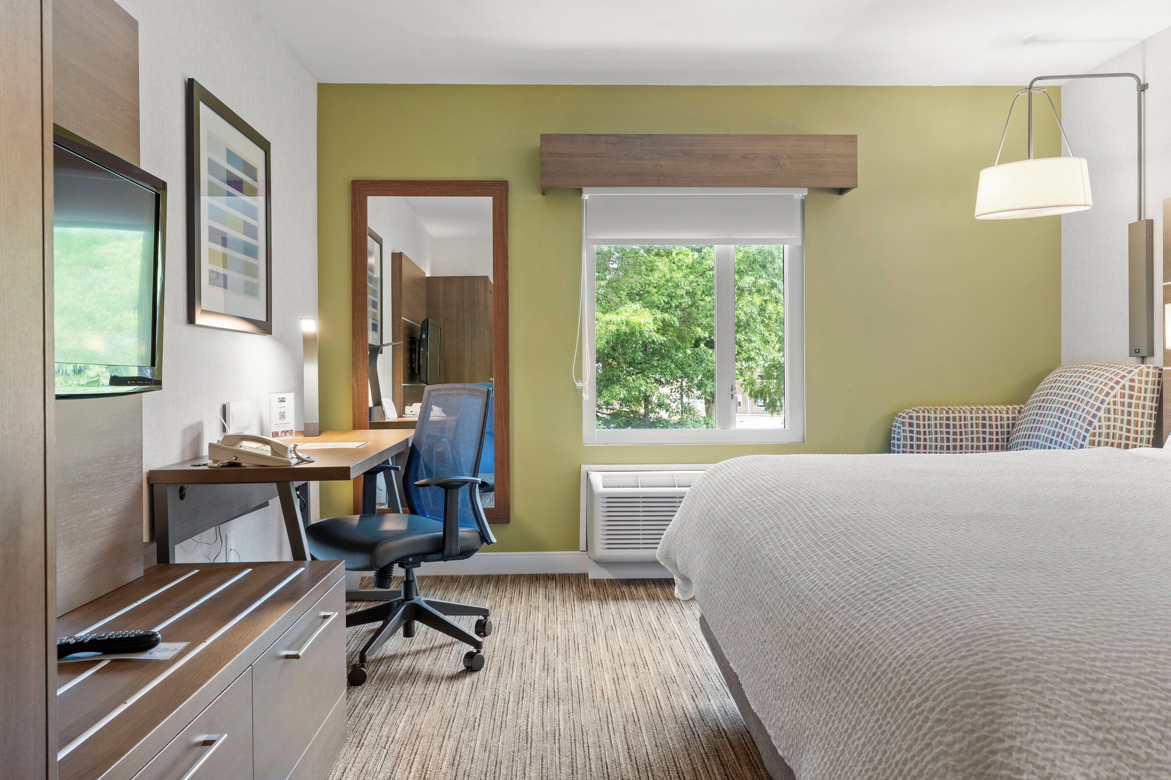A hotel room with a bed, desk, chair, and a large window with a view of trees.