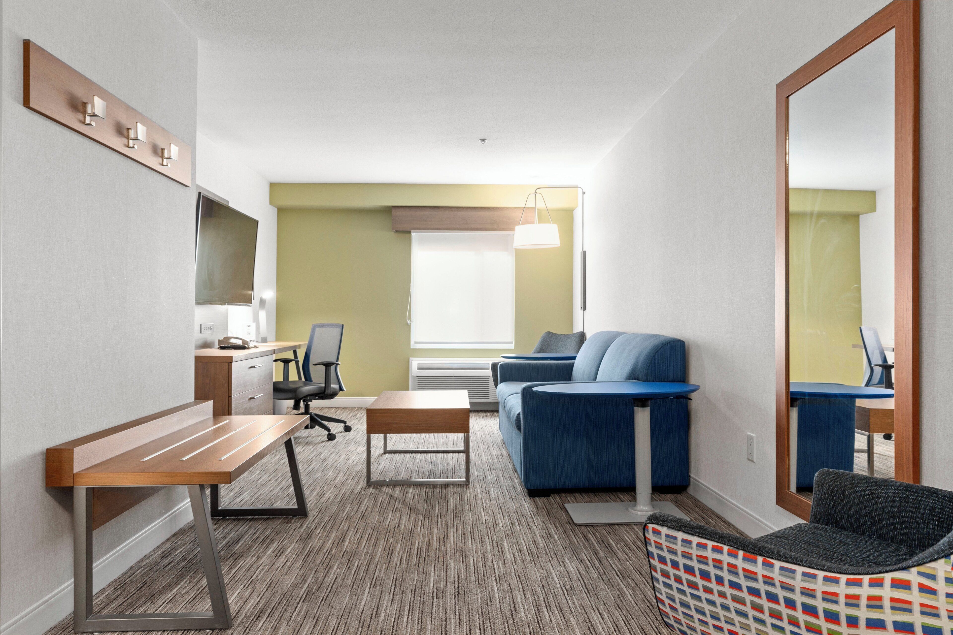 A hotel room with a wooden desk, a blue sofa, a small table, a patterned chair, a large mirror, and a window with curtains.