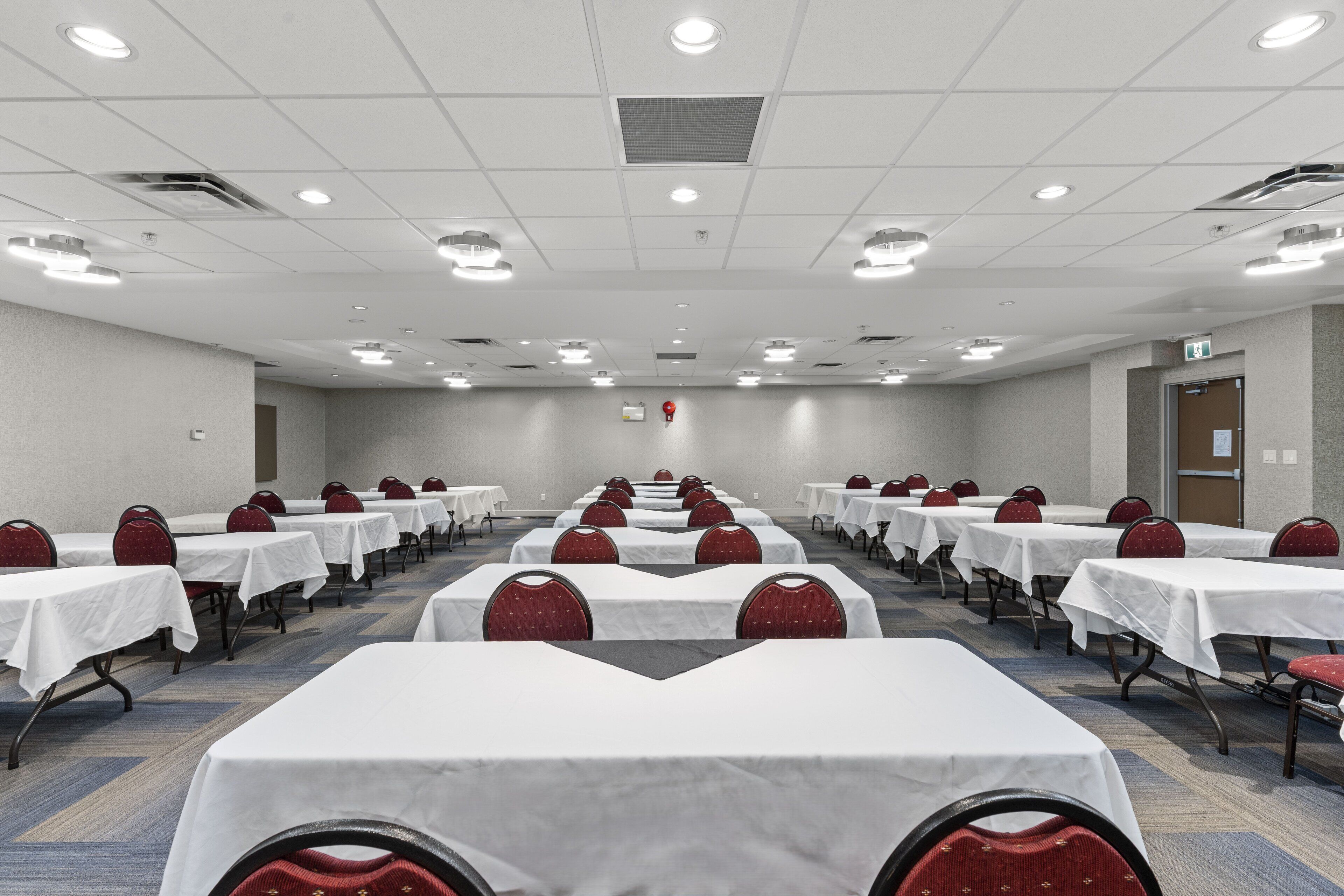 A conference room with rows of tables and chairs, equipped with ceiling lights and a fire extinguisher.
