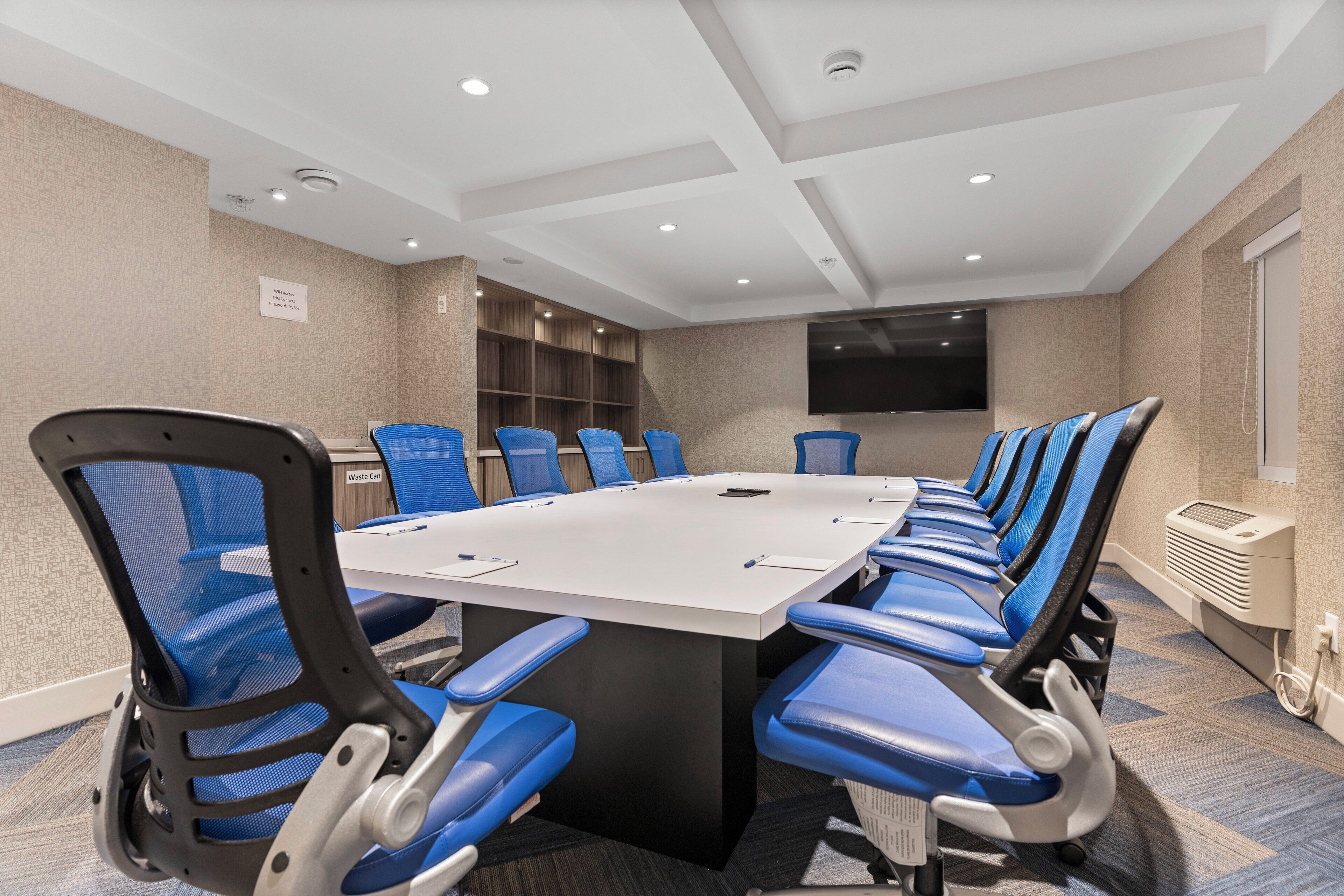 A conference room with a long table, blue chairs, a flat-screen TV, and a waste bin.