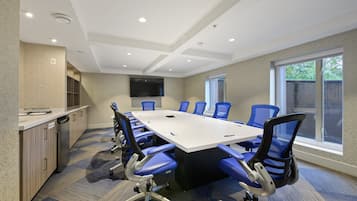 A conference room with a long table, blue chairs, a flat-screen TV, and a kitchen area with a dishwasher.