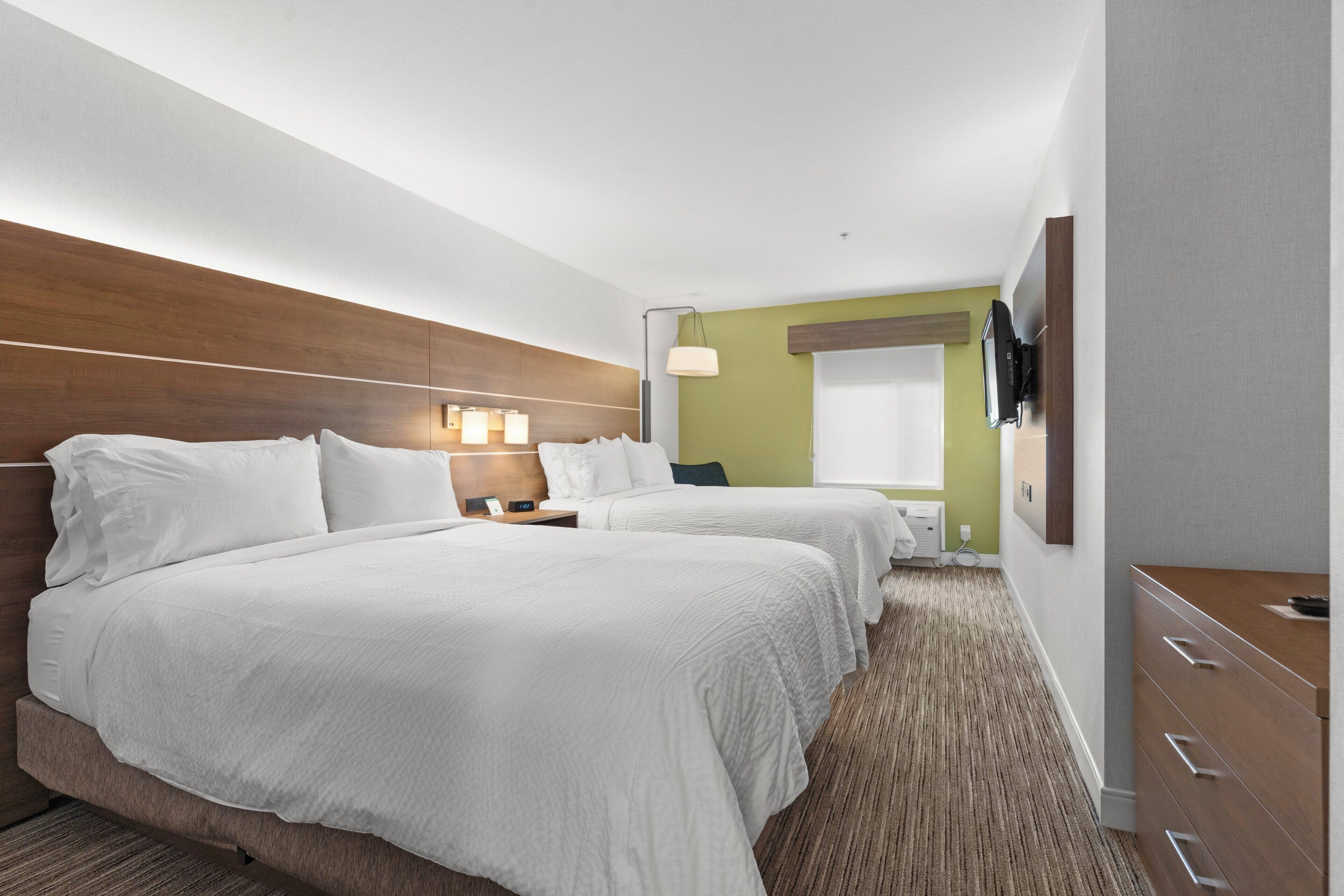 Ein Hotelzimmer mit zwei Betten, einem an der Wand befestigten Fernseher, einem Nachttisch mit Lampe und einem Fenster mit Blick.