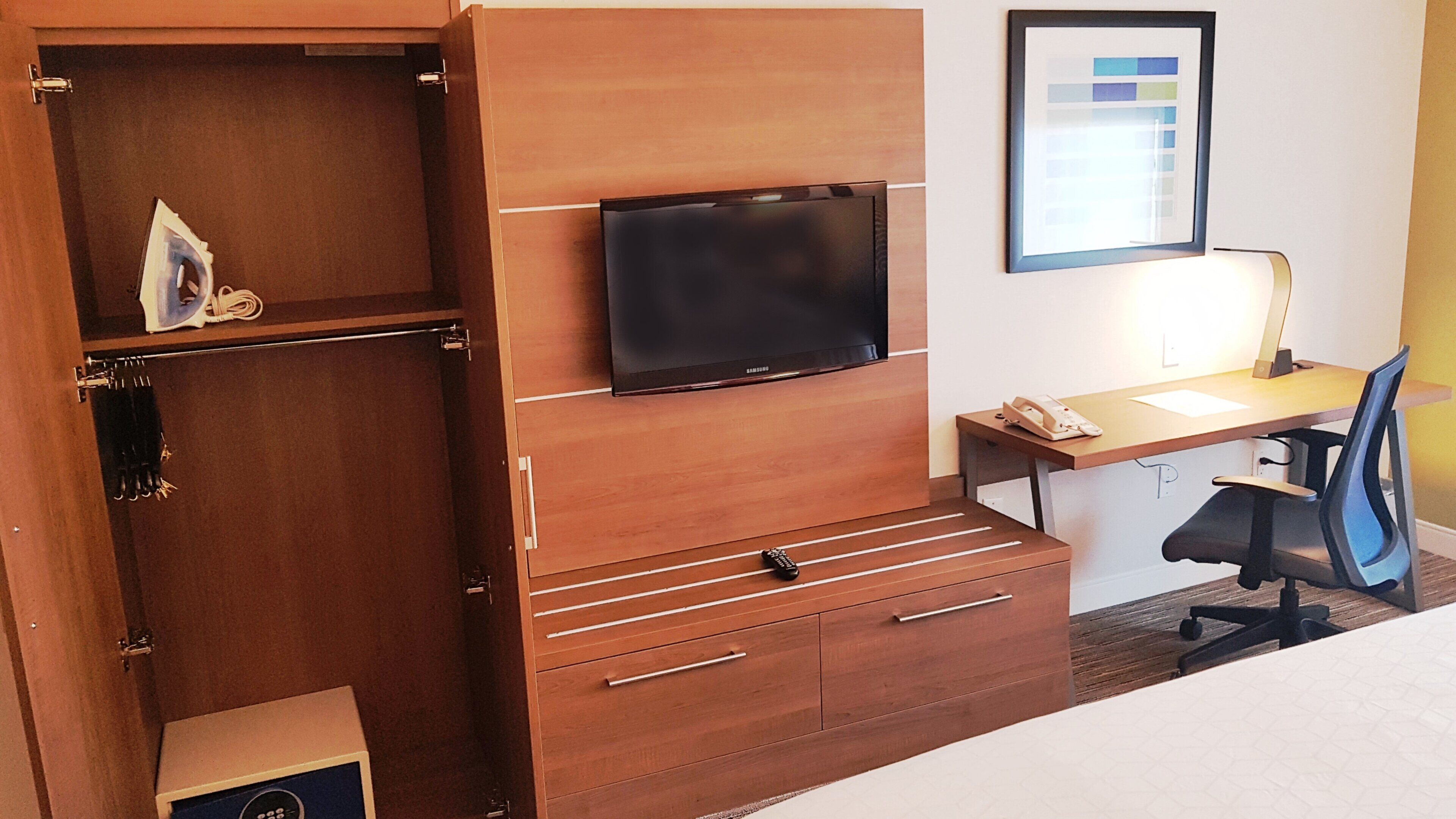Habitación de hotel con cama, escritorio con silla, televisor, armario y un cuadro en la pared.