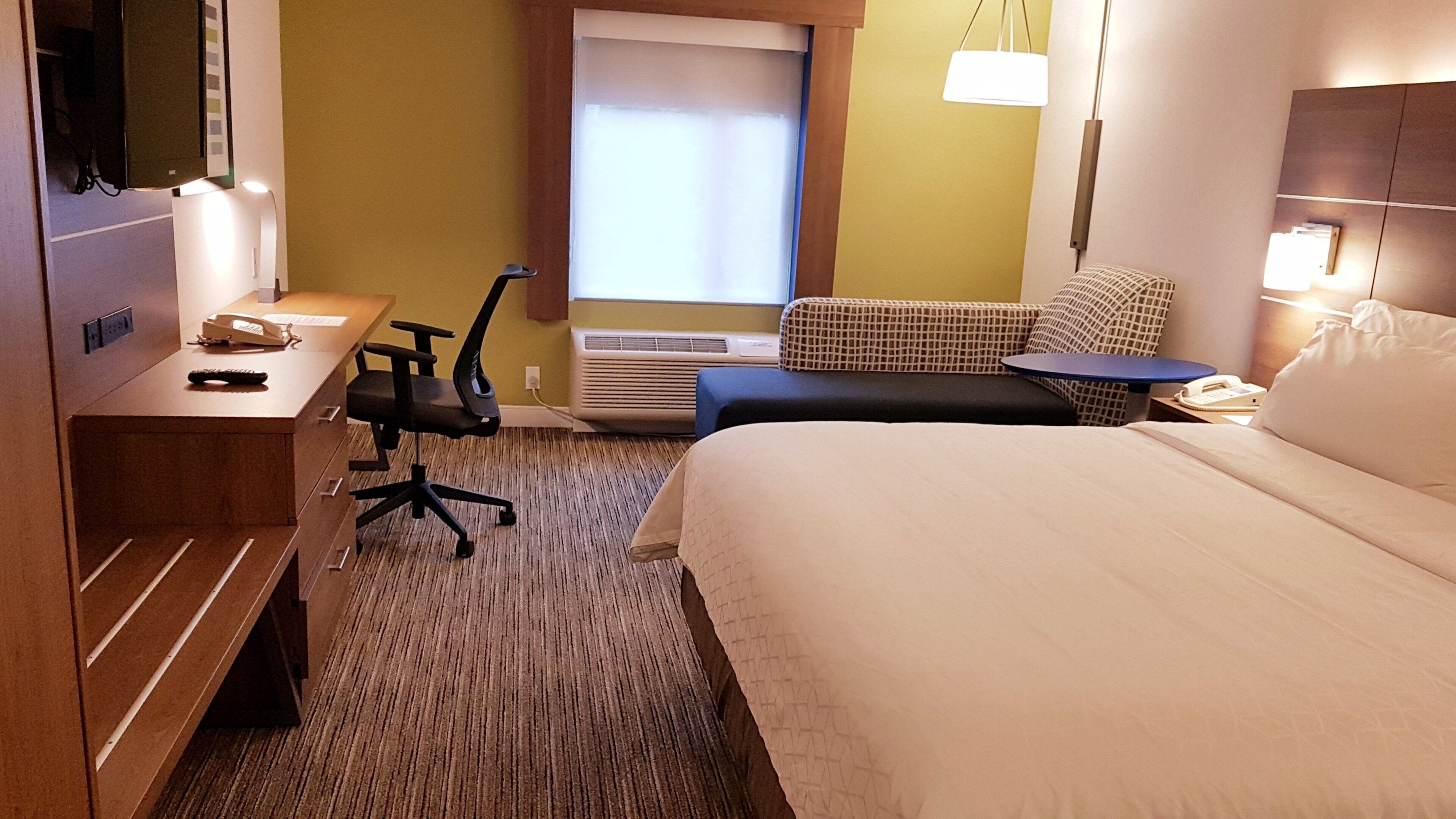 A hotel room with a bed, a desk with a telephone, a chair, a sofa, and a window with blinds.