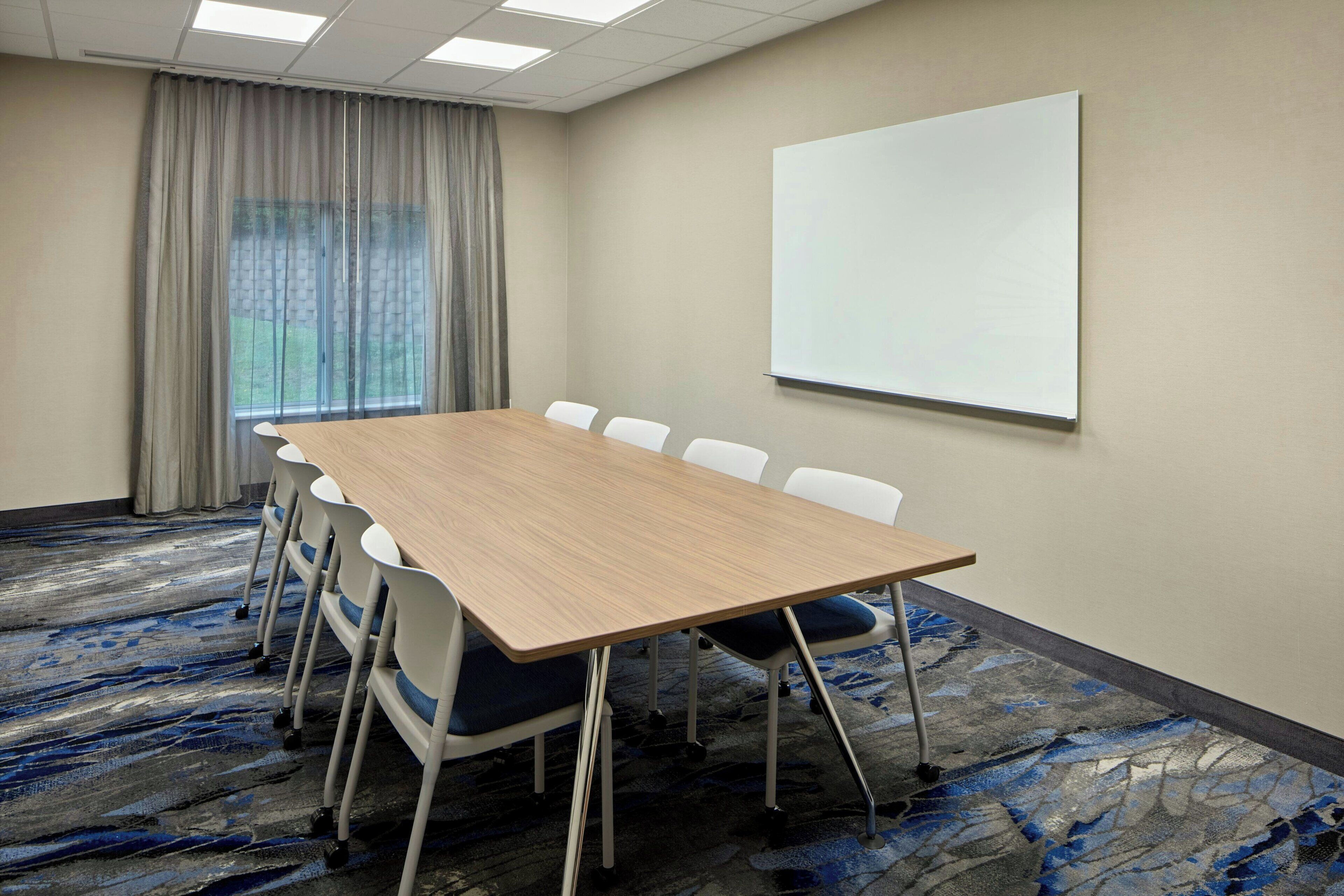 A conference room with a large table, chairs, a projector screen, and a view of the outdoors through a window with curtains.