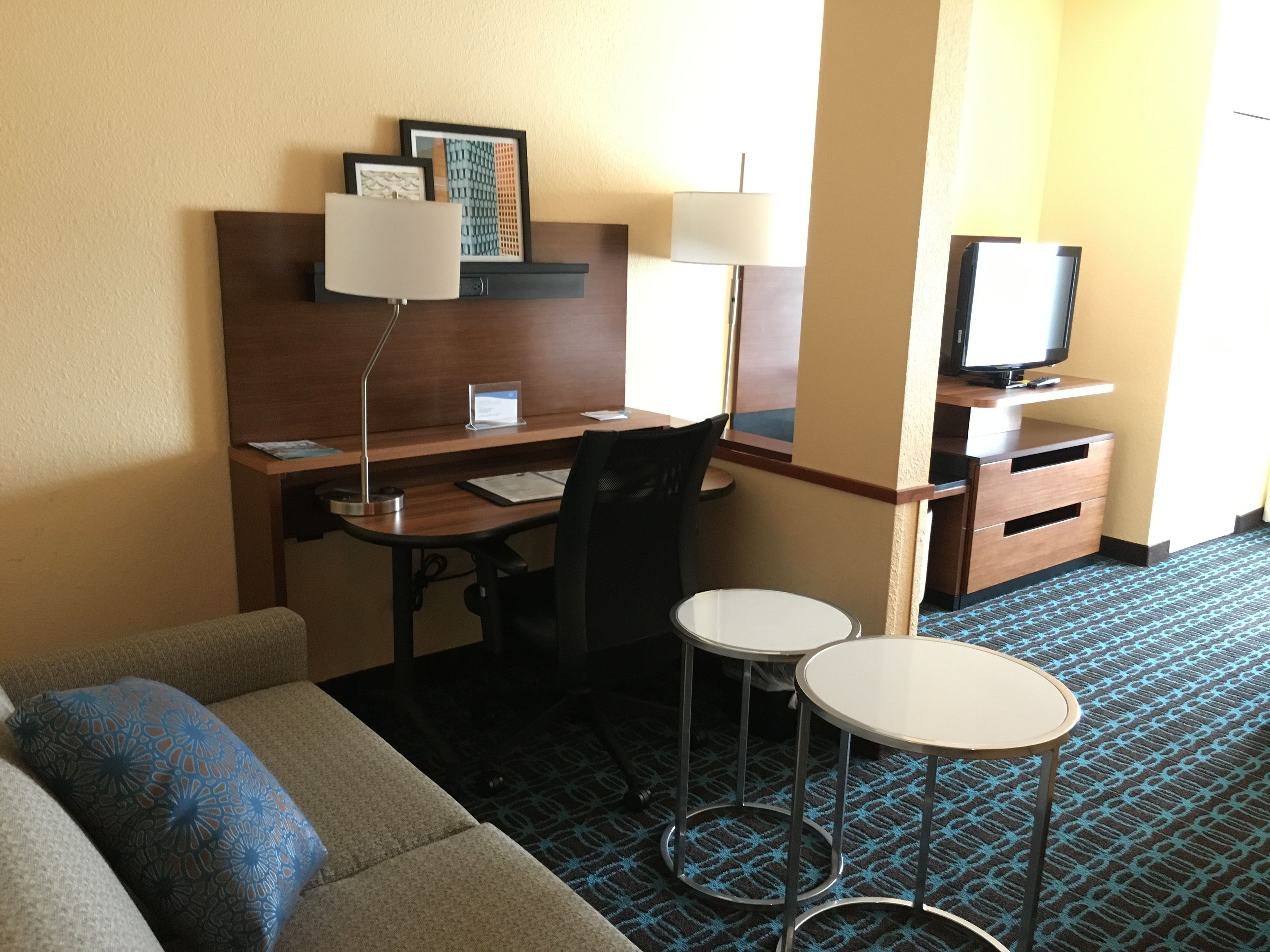 A hotel room with a desk, two chairs, a television, a sofa, and a patterned carpet.