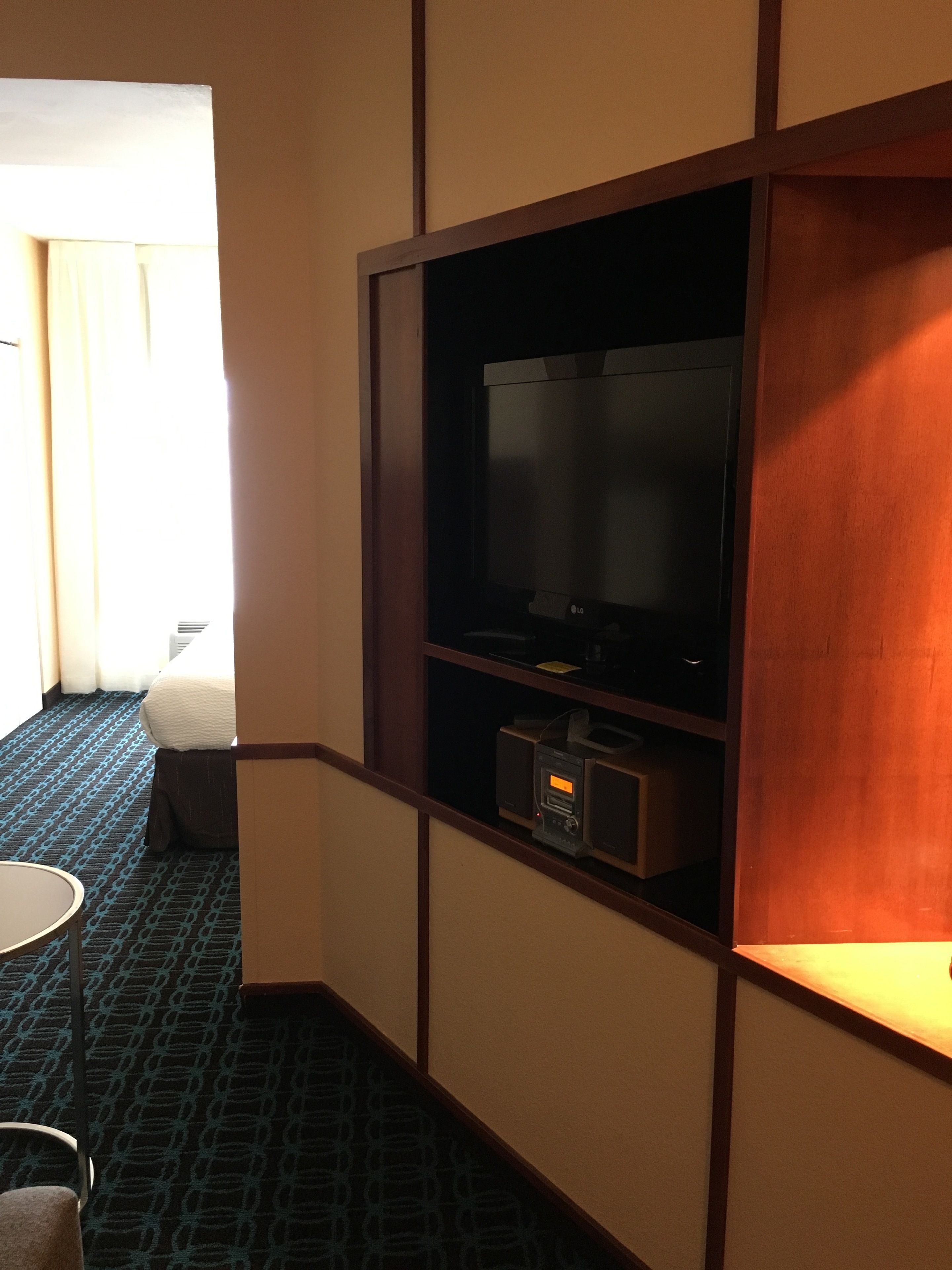 Habitación de hotel con una televisión sobre una estantería de madera, una mesita pequeña y una alfombra estampada.