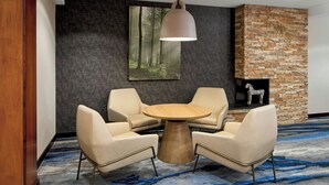 A room with a round wooden table, two beige armchairs, a hanging pendant light, a stone fireplace, and a forest-themed wall art. - Fairfield Inn & Suites by Marriott Frankfort (Frankfort)