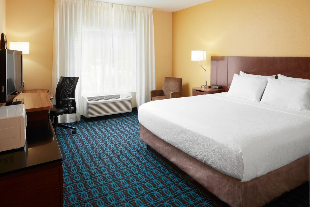 A hotel room with a large bed, a desk with a computer, a chair, a TV, and a window with curtains.