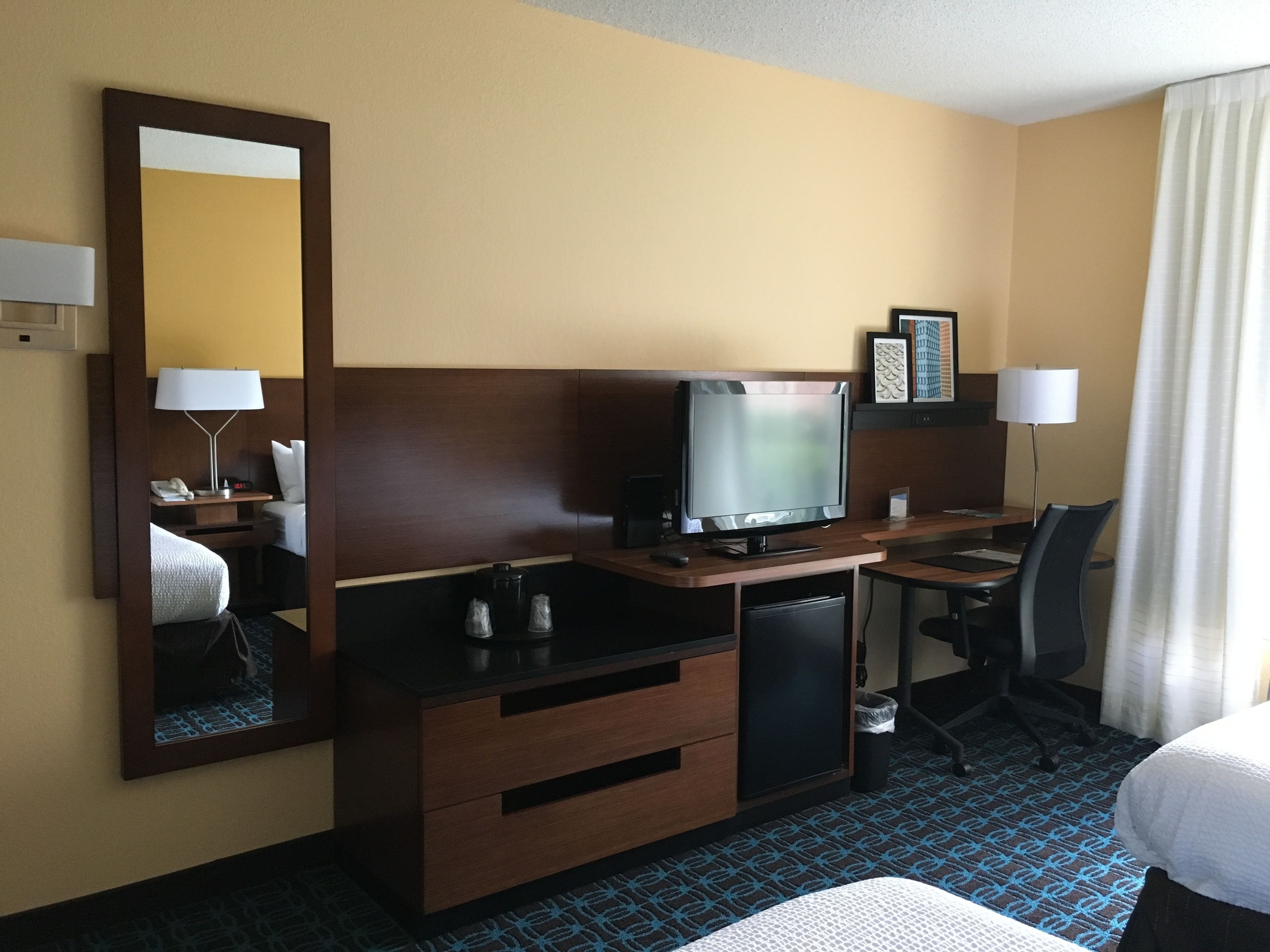 A hotel room with a bed, a desk with a television, a chair, a mirror, and a small refrigerator.