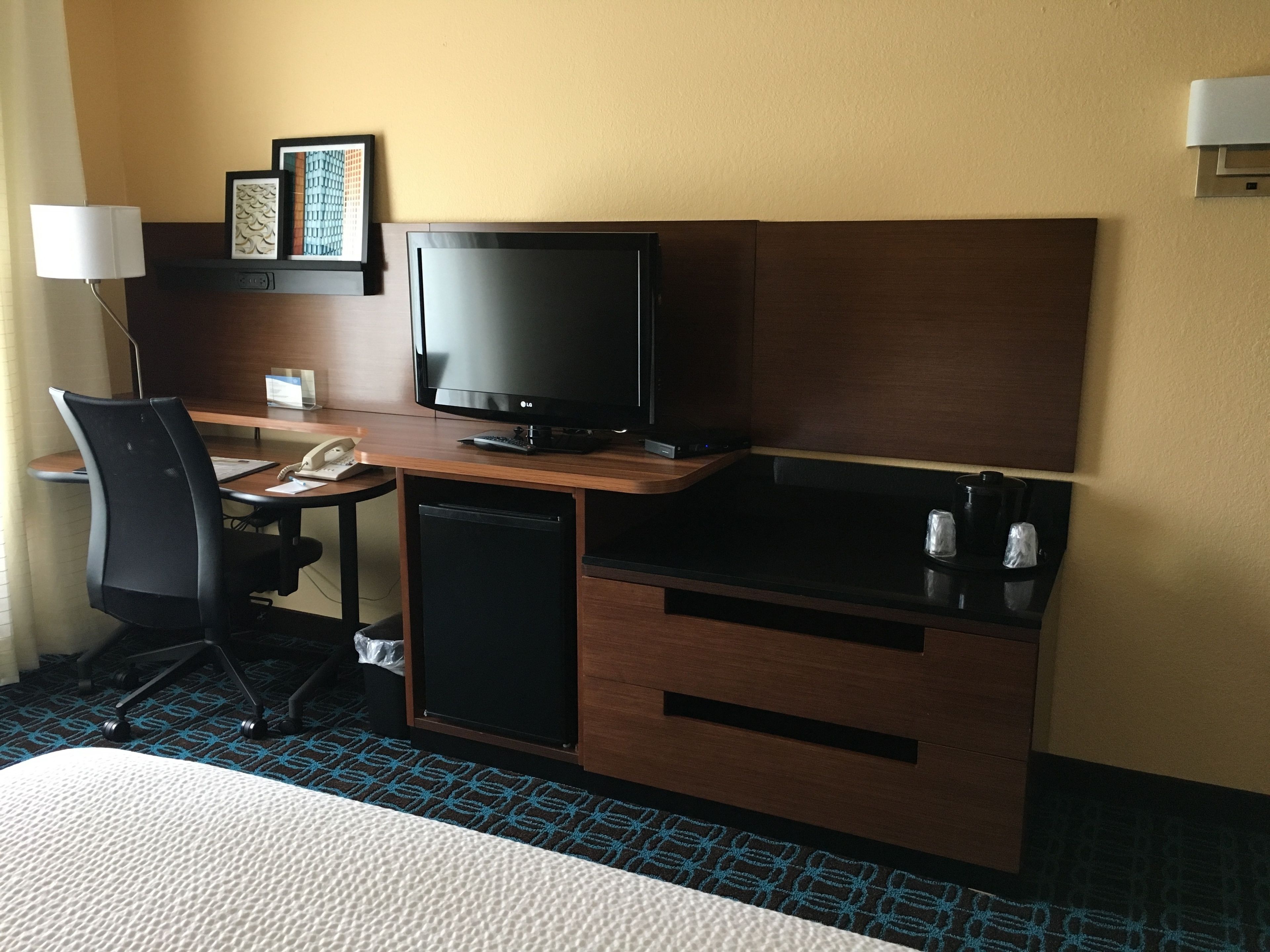 Une chambre d’hôtel avec un bureau en bois, un téléviseur noir, un petit réfrigérateur, une chaise et un lit.
