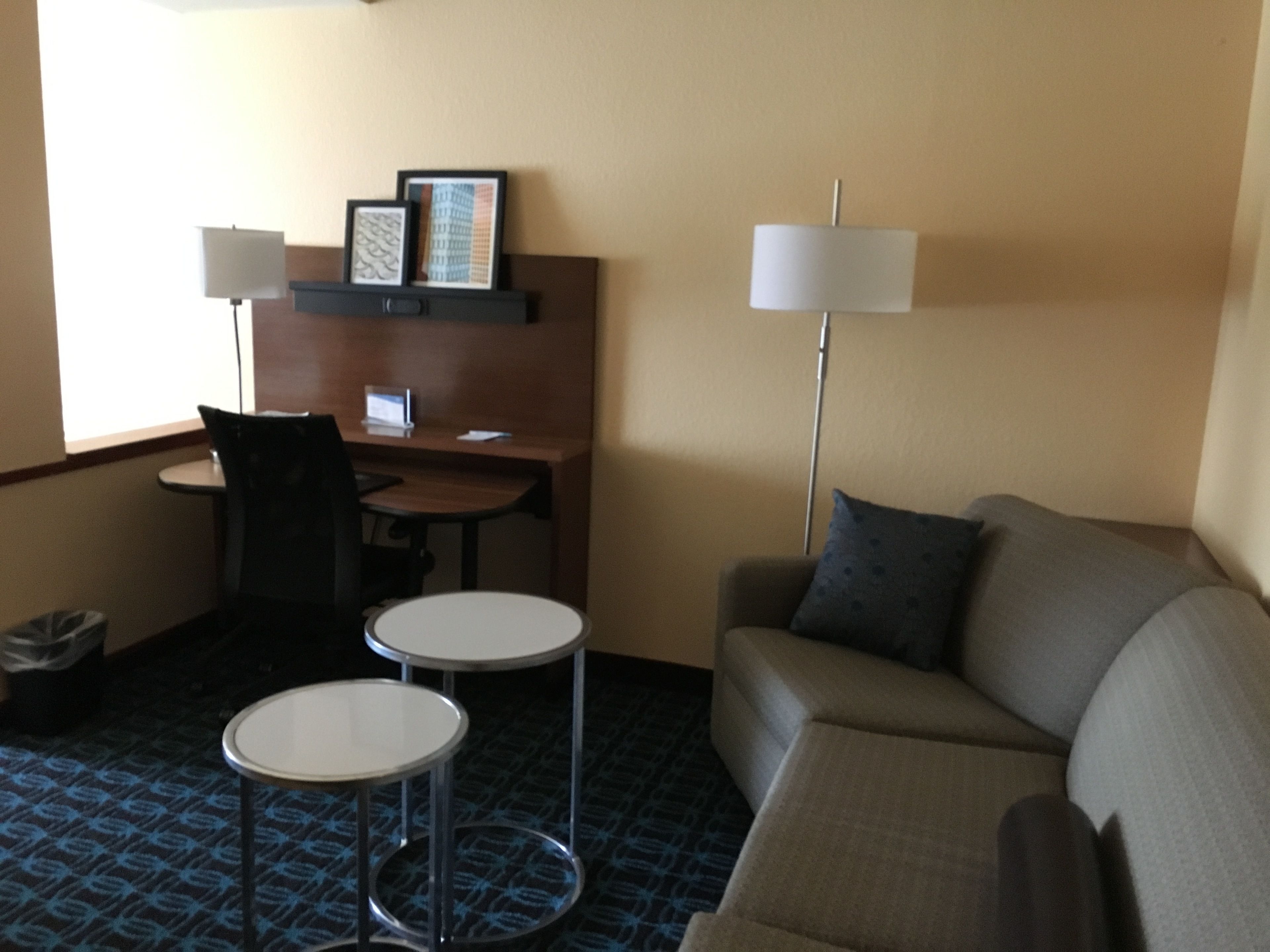Habitación de hotel con sofá, dos mesitas, un escritorio con computadora, lámpara y cuadros en la pared.