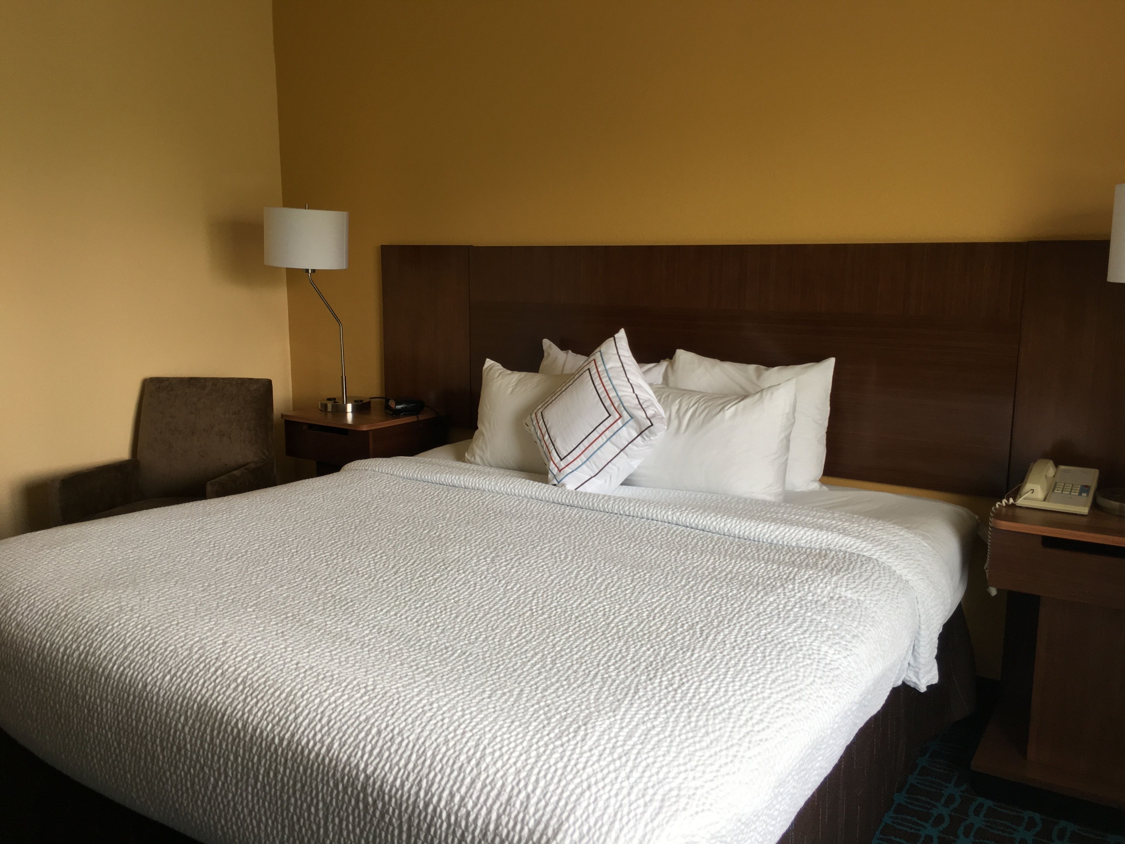 A hotel room with a large bed, a nightstand with a lamp, a chair, and a telephone.