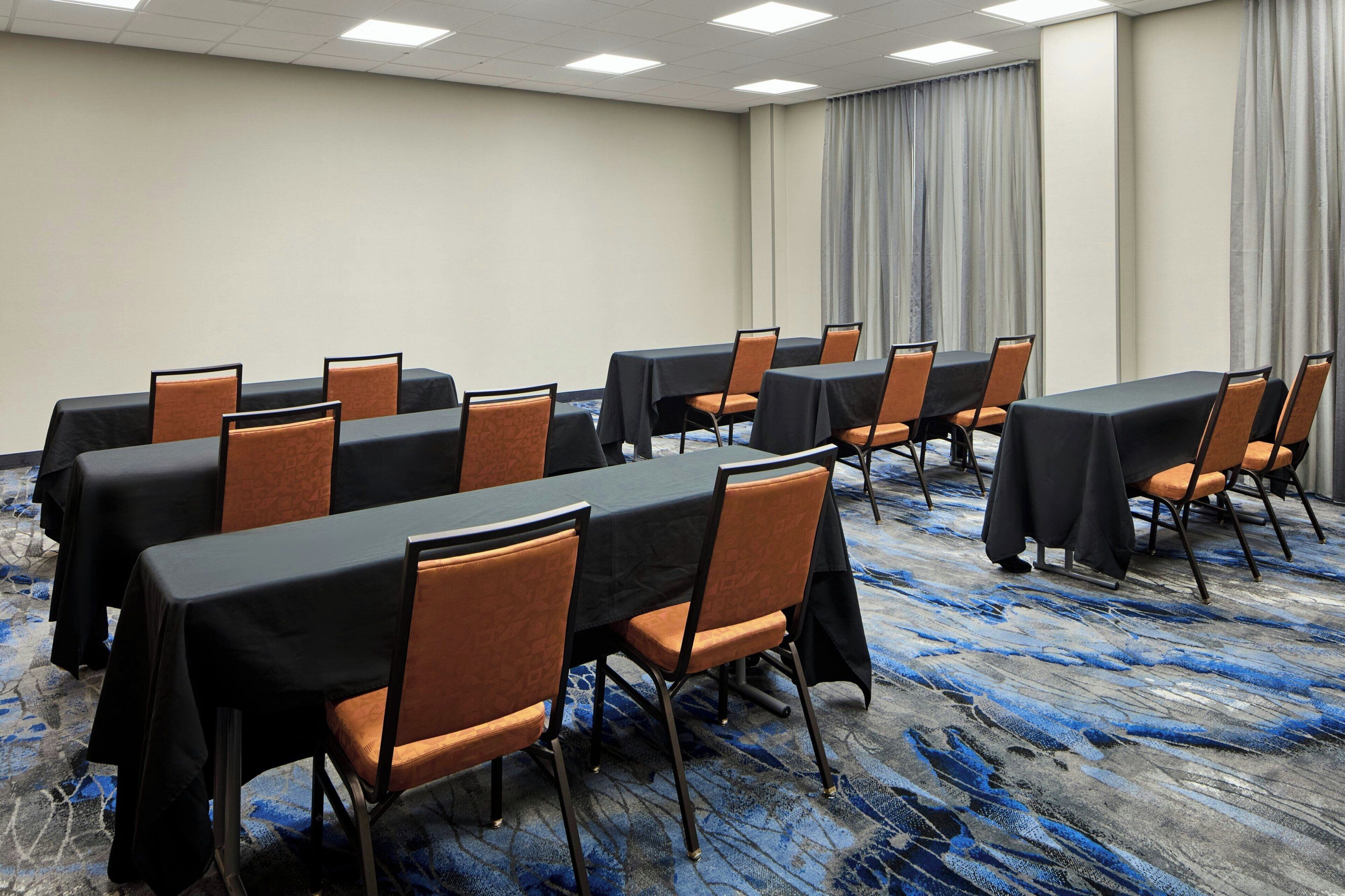 A conference room with tables and chairs, a carpeted floor, and a plain wall with a curtain.