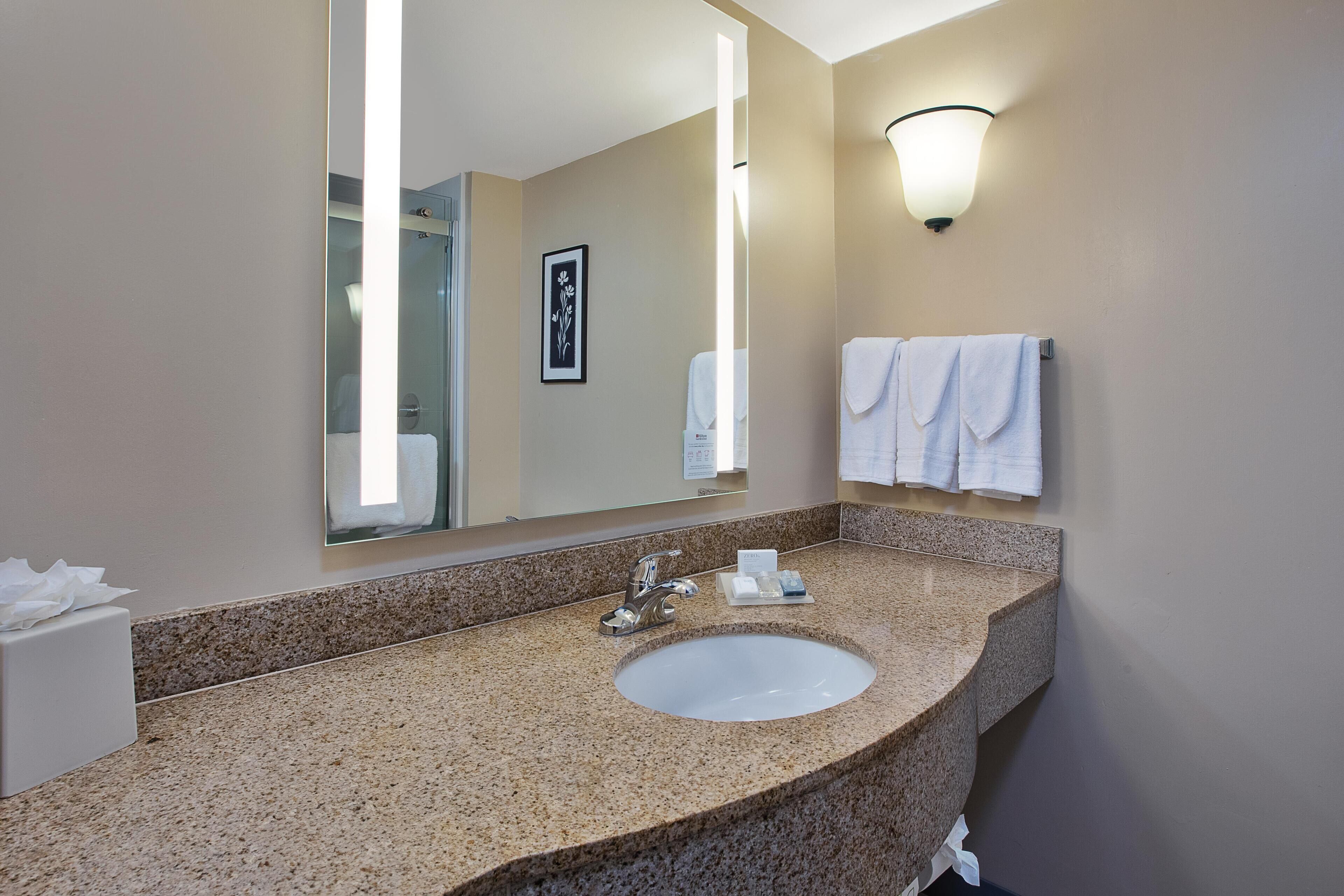 Un baño con encimera de granito, lavamanos blanco, un espejo grande y un toallero de pared.