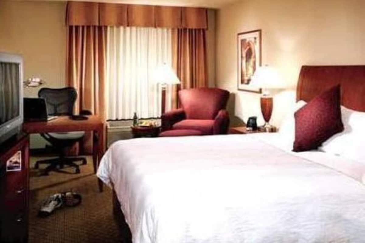A hotel room with a bed, a desk with a computer, a chair, a TV, and a window with curtains.