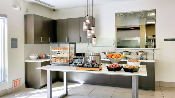 Eine moderne Küche mit Edelstahlgeräten, einer Glastheke voller Gebäck und einer Frühstückstheke mit Früchten und Kaffeeautomaten.