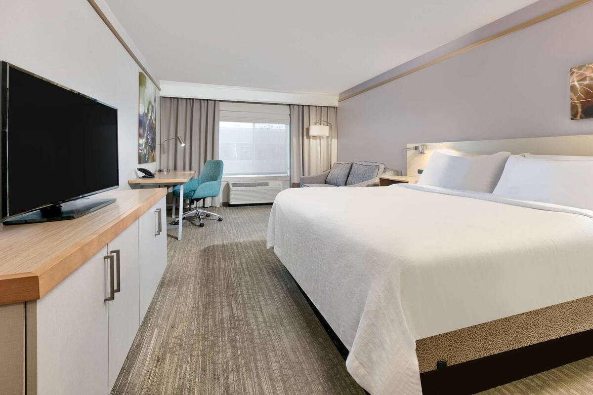A hotel room with a bed, a desk with a TV, a chair, and a window with curtains.