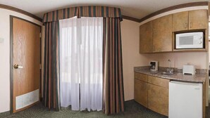 Quarto de hotel com porta de madeira, cortina, cozinha compacta com micro-ondas, máquina de café e pequeno frigorífico.