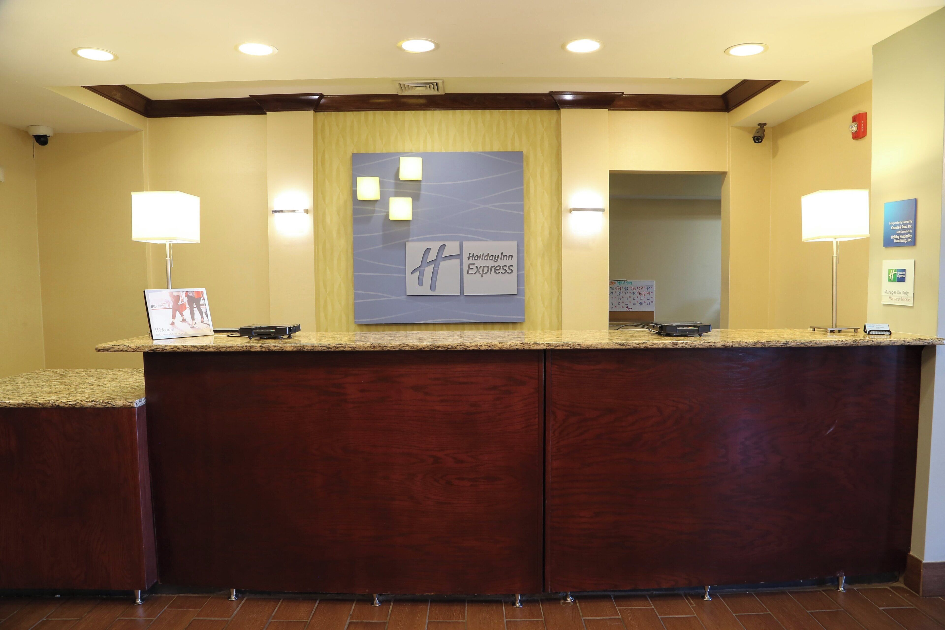Een hotel receptiebalie met een houten aanrecht, een groot bord met Holiday Inn Express erop, en een receptiegedeelte met een spiegel en wandlampen.