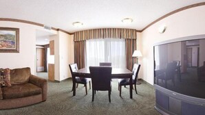 Quarto de hotel com mesa de jantar, quatro cadeiras, sofá, televisão e uma janela grande com cortinas.