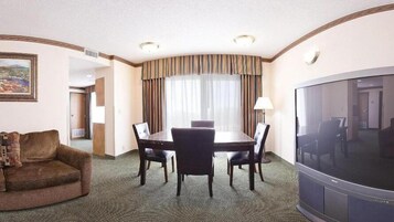 Habitación de hotel con una mesa de comedor, cuatro sillas, un sofá, un televisor y un ventanal con cortinas.