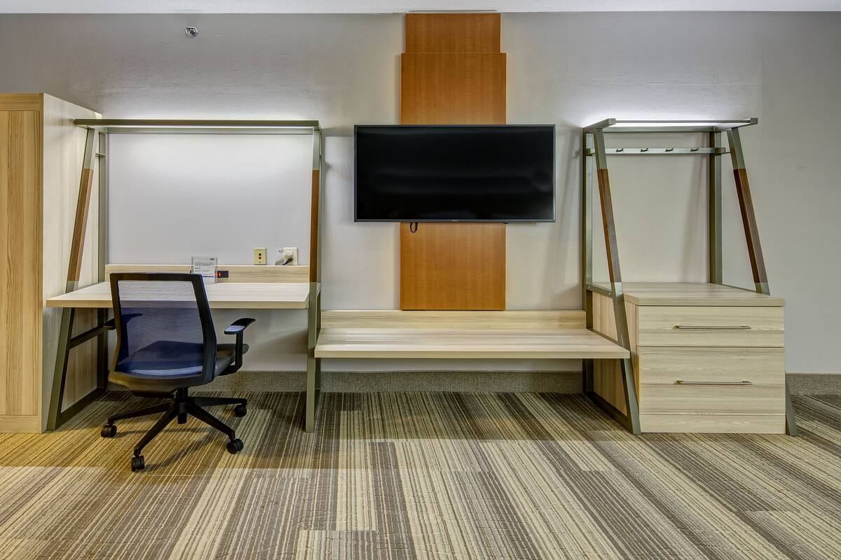 A room with a wooden wardrobe, a desk with a chair, a TV mounted on the wall, and a small cabinet.