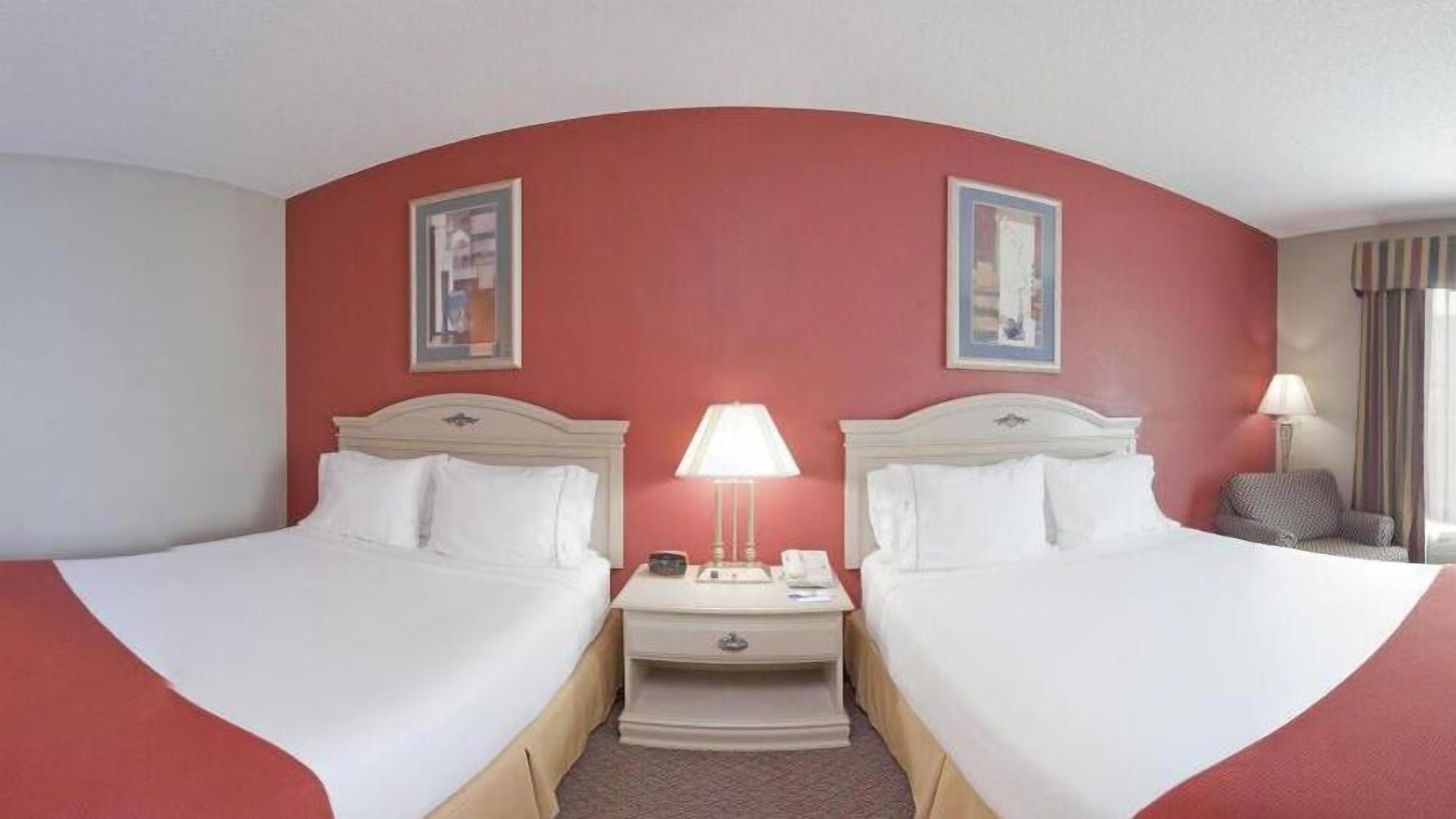 Quarto de hotel com duas camas, uma mesa de cabeceira com luminária e uma cadeira.