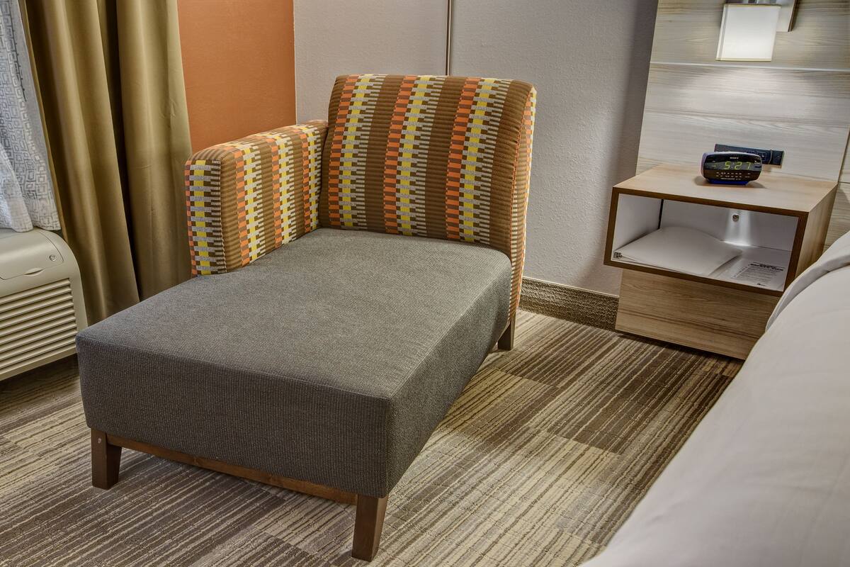 A hotel room with a bed, a small bedside table, a wall-mounted air conditioner, and a window with curtains.