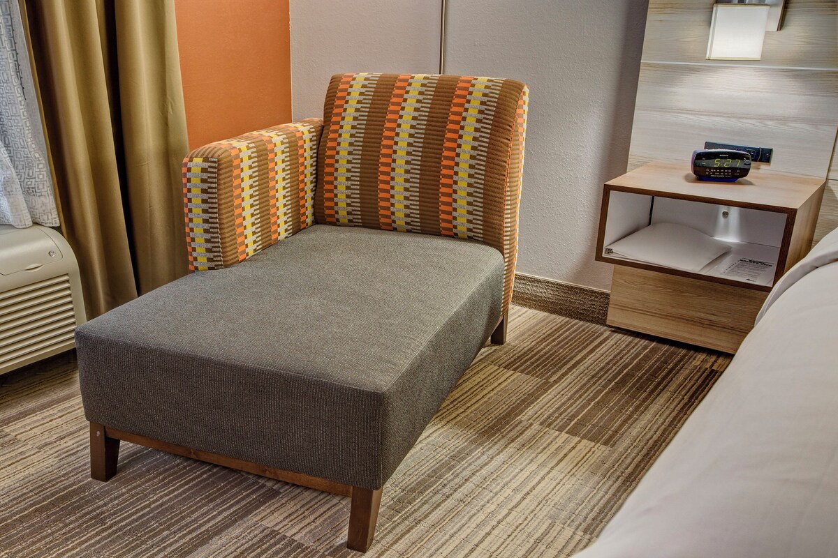 A hotel room with a bed, a nightstand, a wall clock, and a window with curtains.