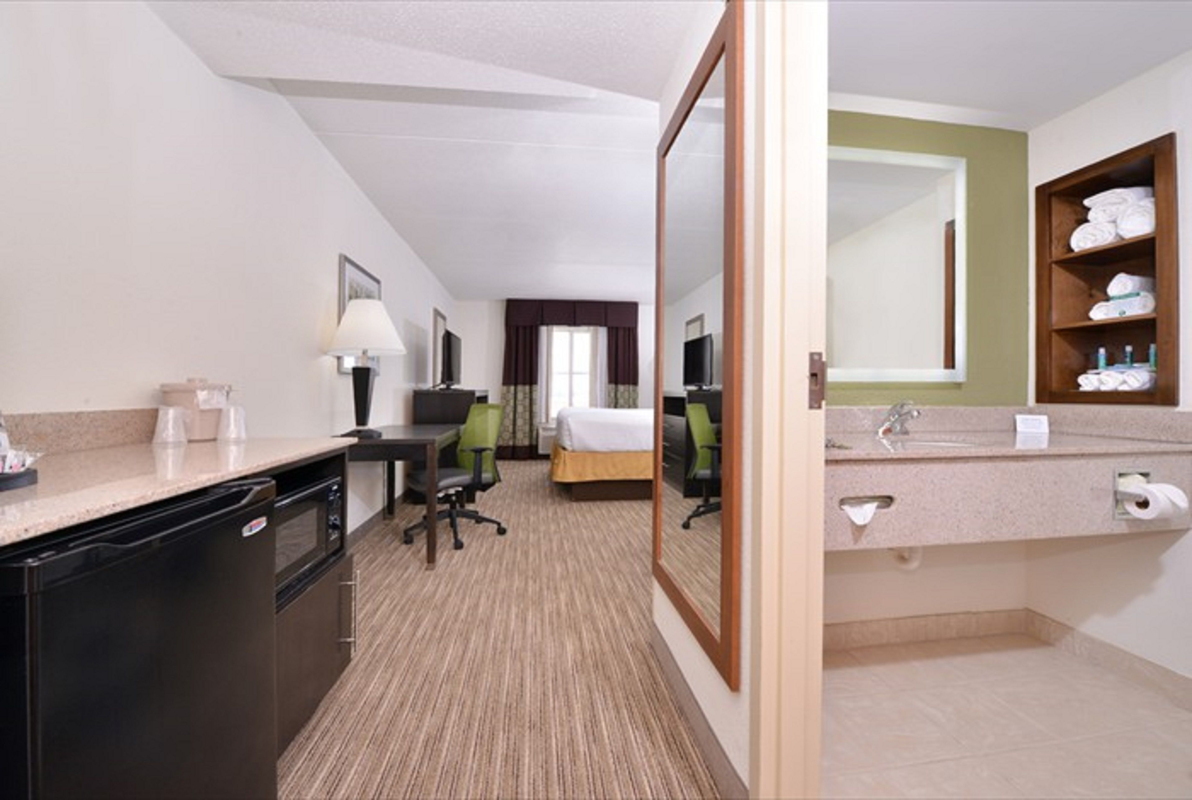 Habitación de hotel con kitchenette, cama, escritorio, silla, espejo y baño con lavabo y toallero.