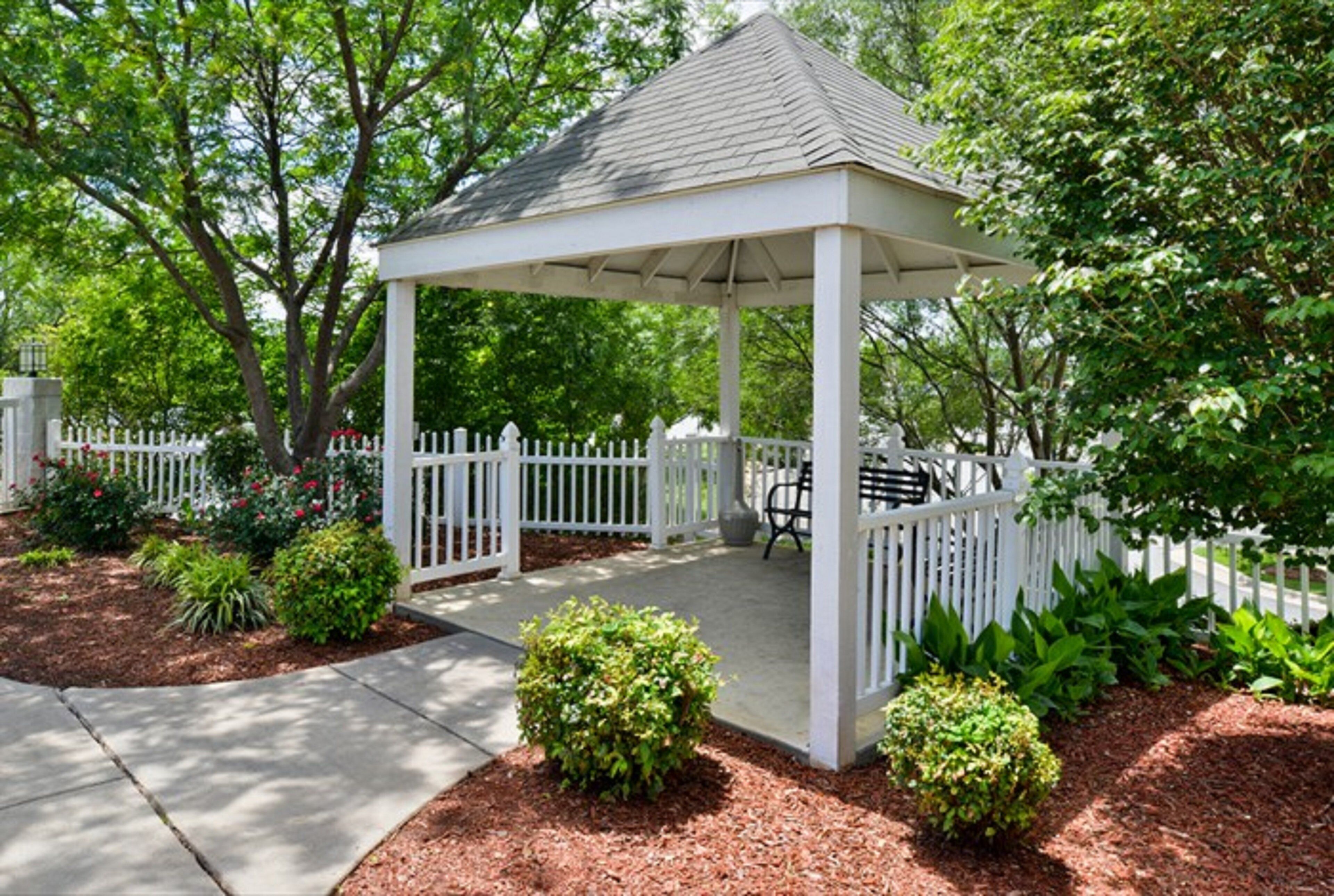 A gazebo with a white roof and columns, surrounded by a white picket fence, set in a landscaped garden with mulch and small bushes.
