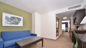Habitación de hotel con sofá azul, una mesa de madera, kitchenette y un cuadro abstracto enmarcado en la pared.
