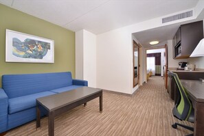 Habitación de hotel con sofá azul, una mesa de madera, kitchenette y un cuadro abstracto enmarcado en la pared.
