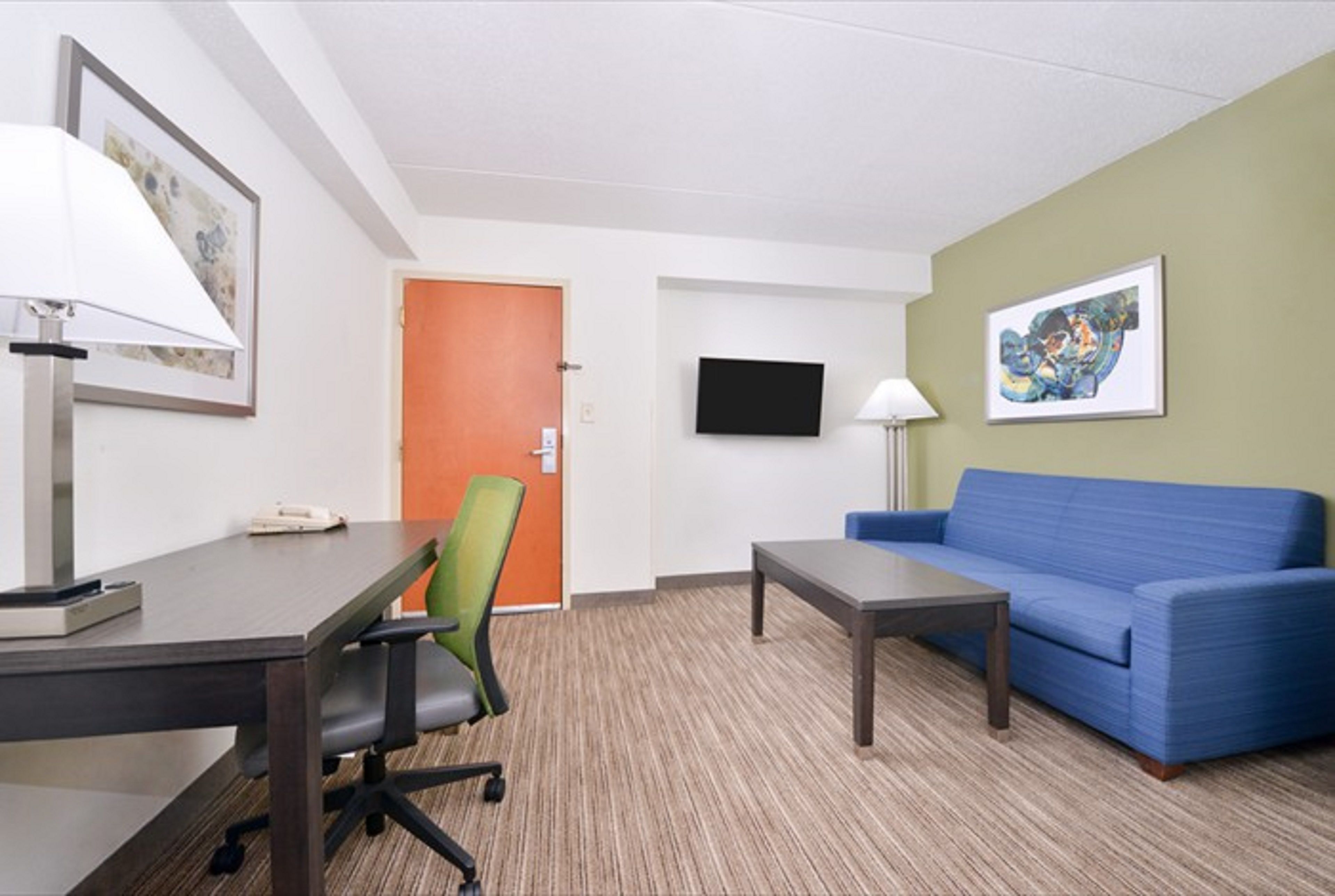 A hotel room with a desk, chair, sofa, TV, and artwork on the walls.